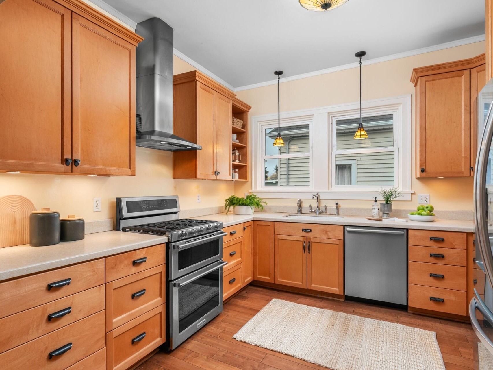 A modern kitchen with wooden cabinets, stainless steel appliances, and light-colored countertops. Two pendant lights hang above the sink. The room is bright with natural light from two windows above the sink. A rug is on the wooden floor.