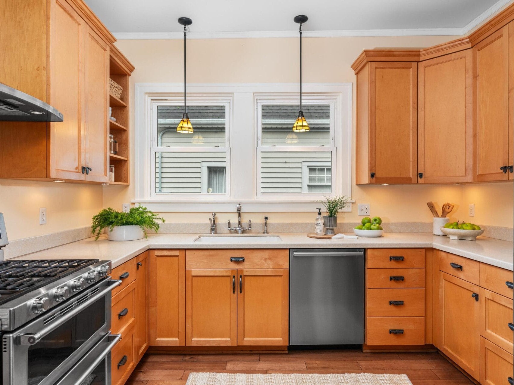 A cozy kitchen with wooden cabinets, stainless steel appliances, and a double sink under two windows. Pendant lights hang from the ceiling, and a green plant with a bowl of apples adds a touch of freshness to the counter.