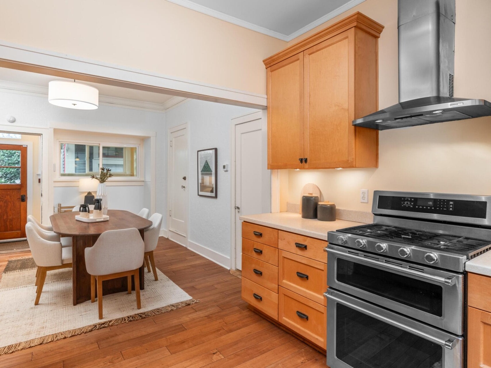 A modern kitchen and dining area with wooden cabinets, a stainless steel oven, and a range hood. Adjacent is a dining table with six beige chairs. A door and a window add natural light to the space, highlighting the polished wooden floor.