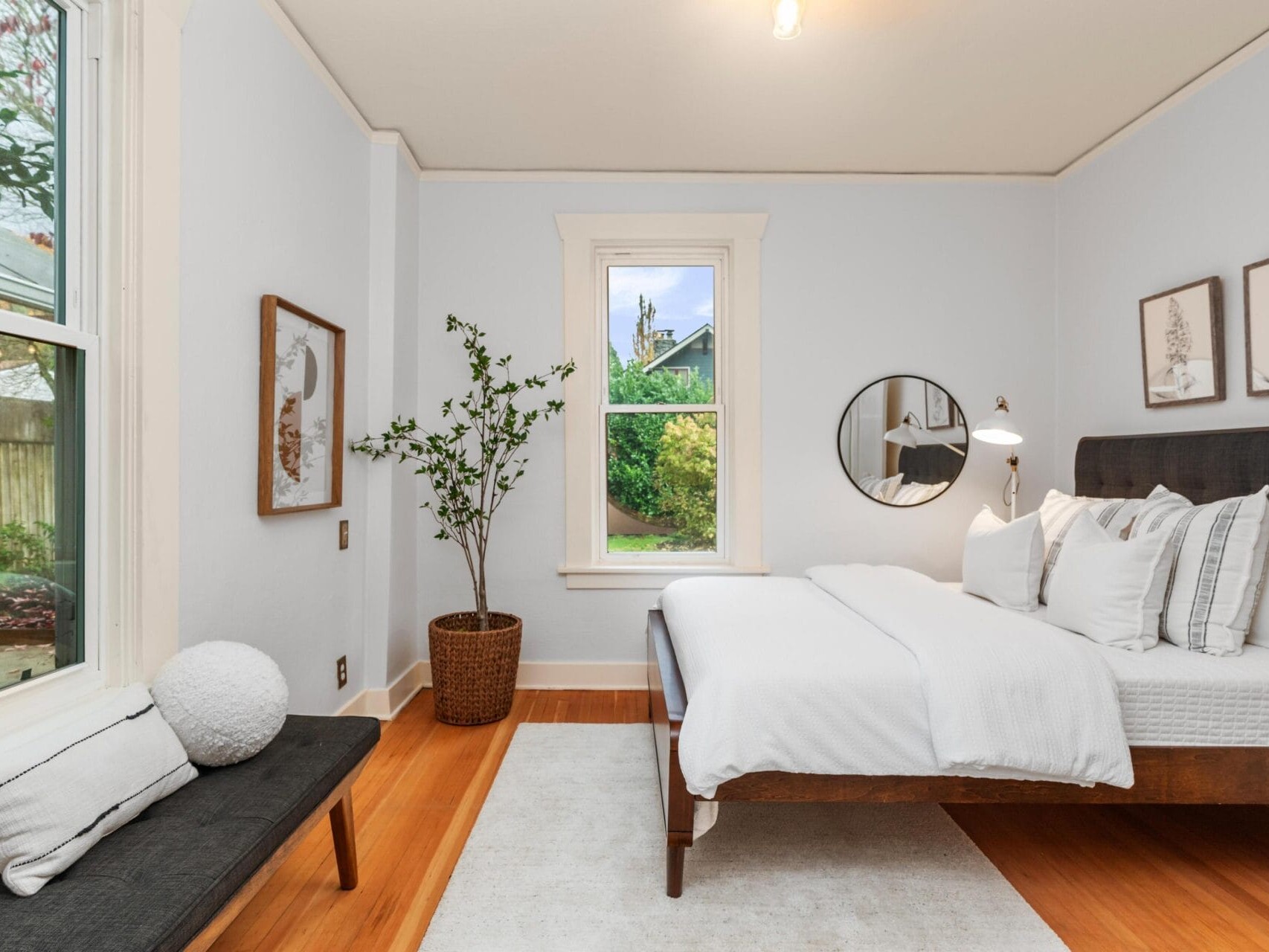 A bright, minimalist bedroom with a large bed featuring white linens and gray accent pillows. A black bench with a white cushion sits by a window. Theres a round mirror, framed art on the walls, and a potted plant accentuates the space.
