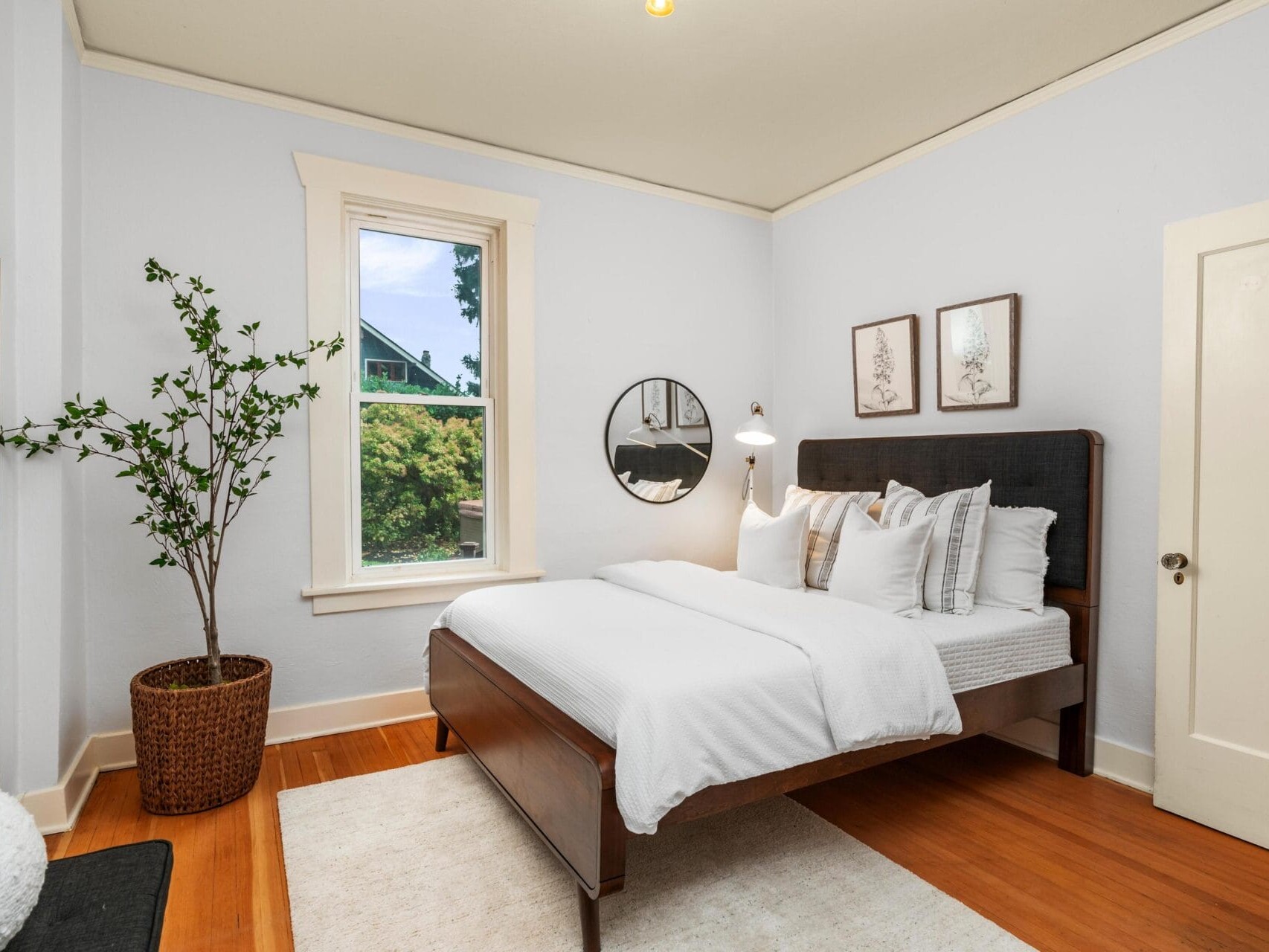 A cozy, light-filled bedroom with a neatly made double bed featuring white bedding and multiple pillows. The room has a potted plant, a round mirror, and framed art on the walls, with a window showcasing greenery outside.