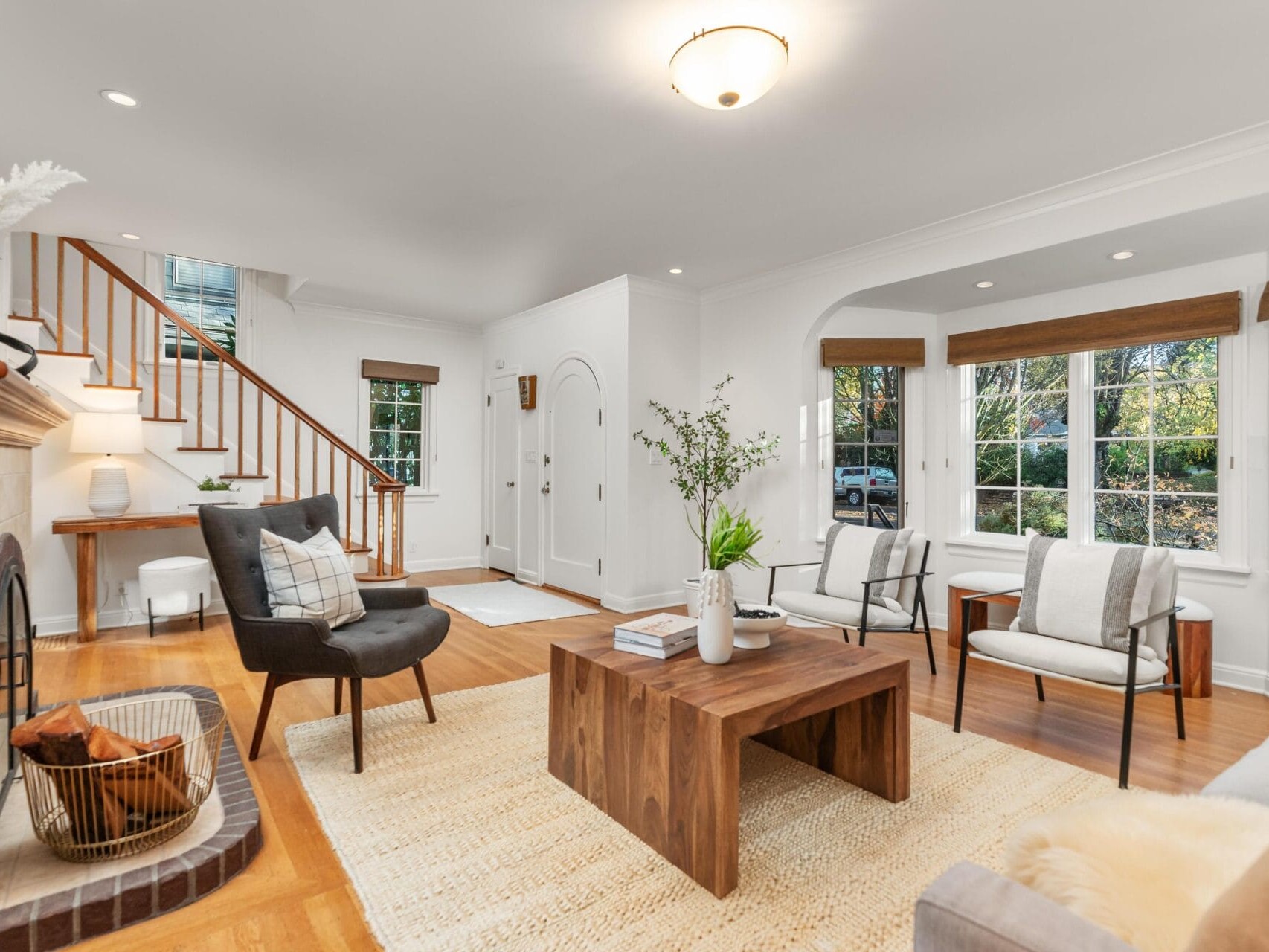 A cozy living room with wooden floors and a neutral color scheme. Features include a wooden coffee table, armchairs, a fireplace, and a basket by the hearth. Large windows with brown blinds let in natural light, and a staircase leads to the upper floor.