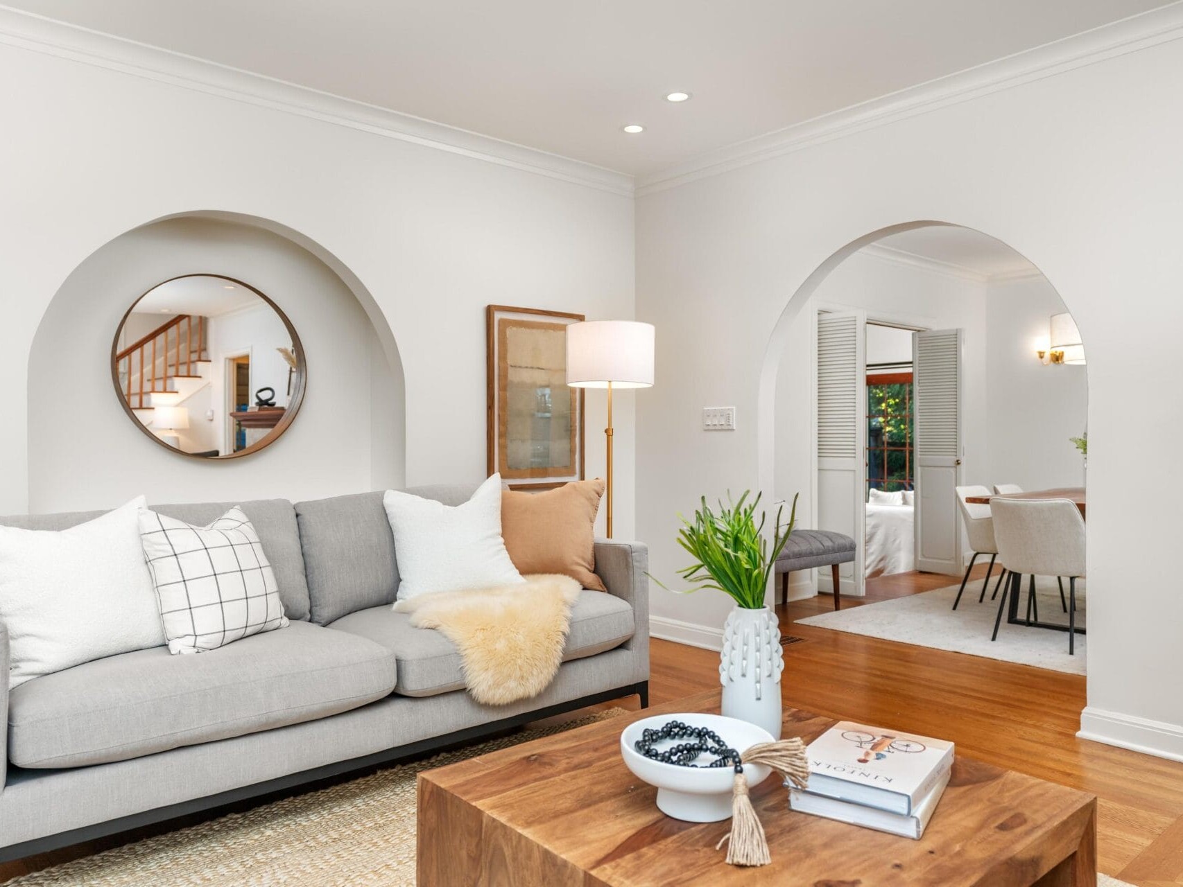 A modern living room with a gray sofa decorated with white and beige cushions, a wooden coffee table with books and decor, and arched doorways. A round mirror and floor lamp adorn the space, leading to a dining area with a wooden table and chairs.