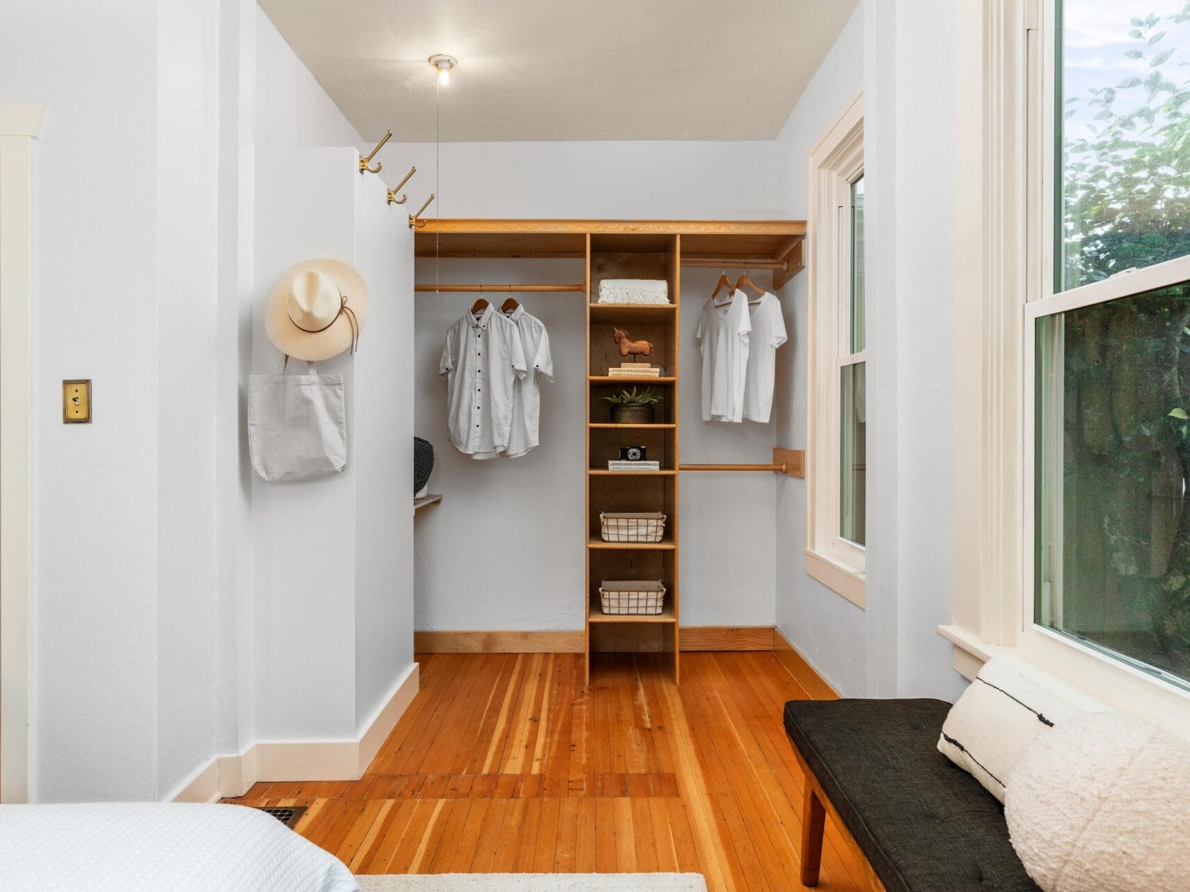 A minimalist walk-in closet with wooden shelves and a hanging rod. It contains white shirts, a hat, a light bag, towels, and a small plant. The room has white walls, wooden flooring, and a window with a bench underneath.