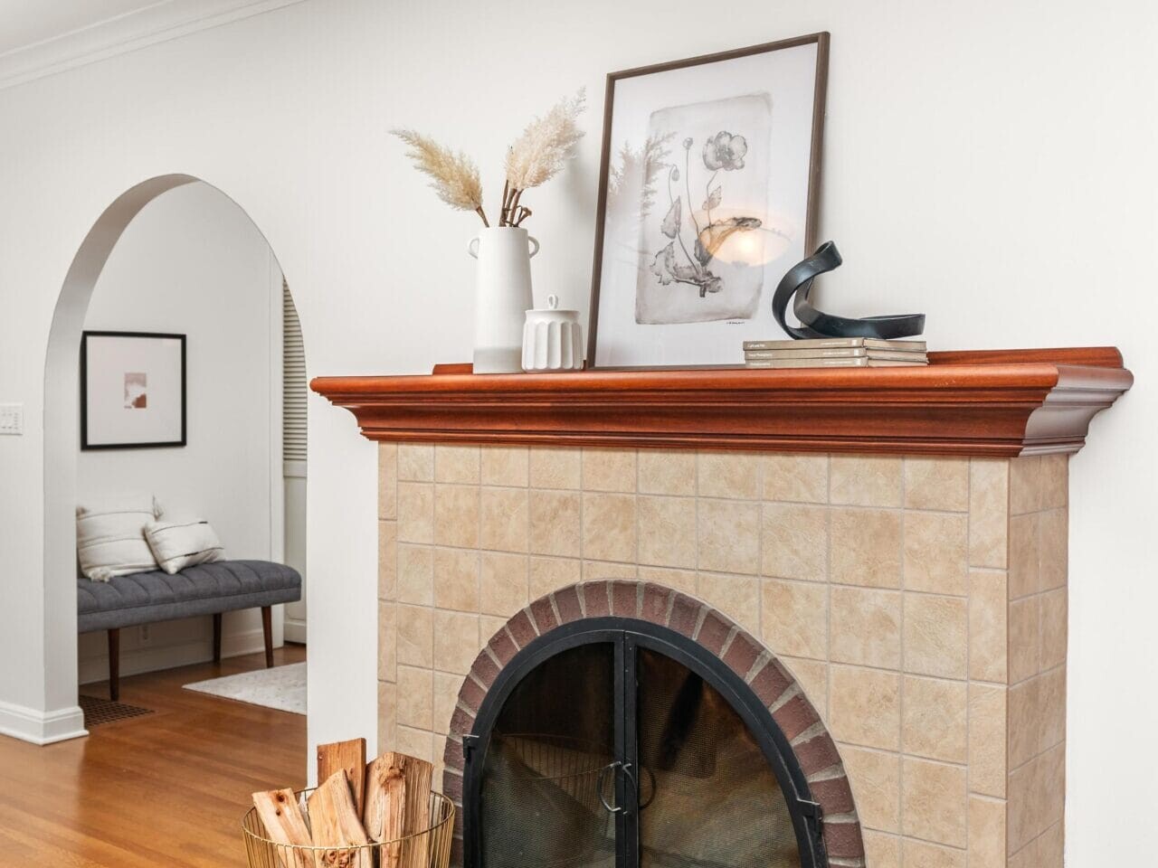 A cozy living room features a tiled fireplace with a wooden mantel, adorned with decorative items and artwork. A basket of firewood sits nearby. An archway leads to a seating area with a bench, all set on polished hardwood floors.