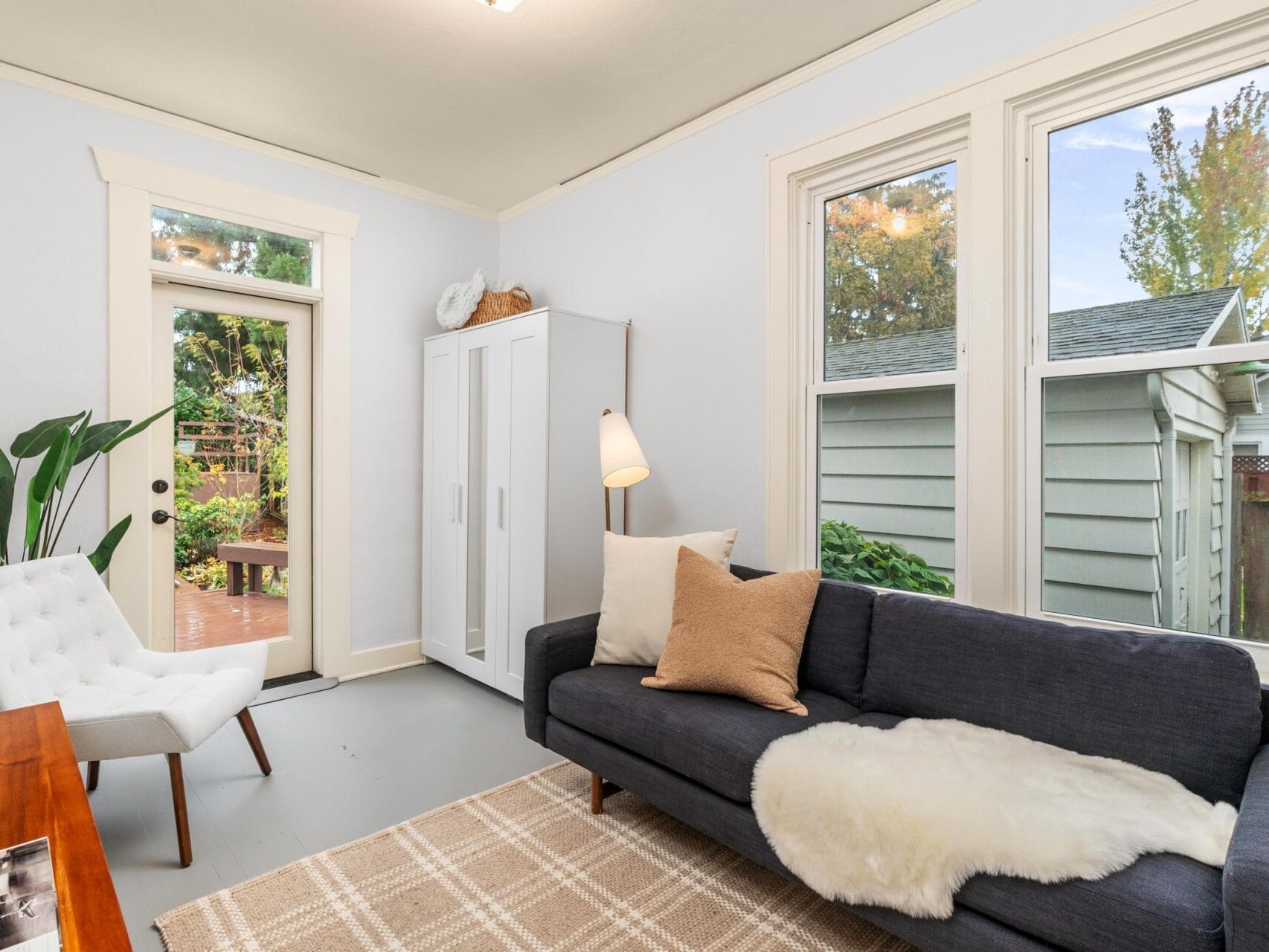 A cozy living room with a dark sofa, white chair, and a plaid rug. A fluffy throw is on the sofa, and a tall plant sits near the door, which leads to a backyard. Large windows offer a view of the garden and a white wardrobe stands in the corner.