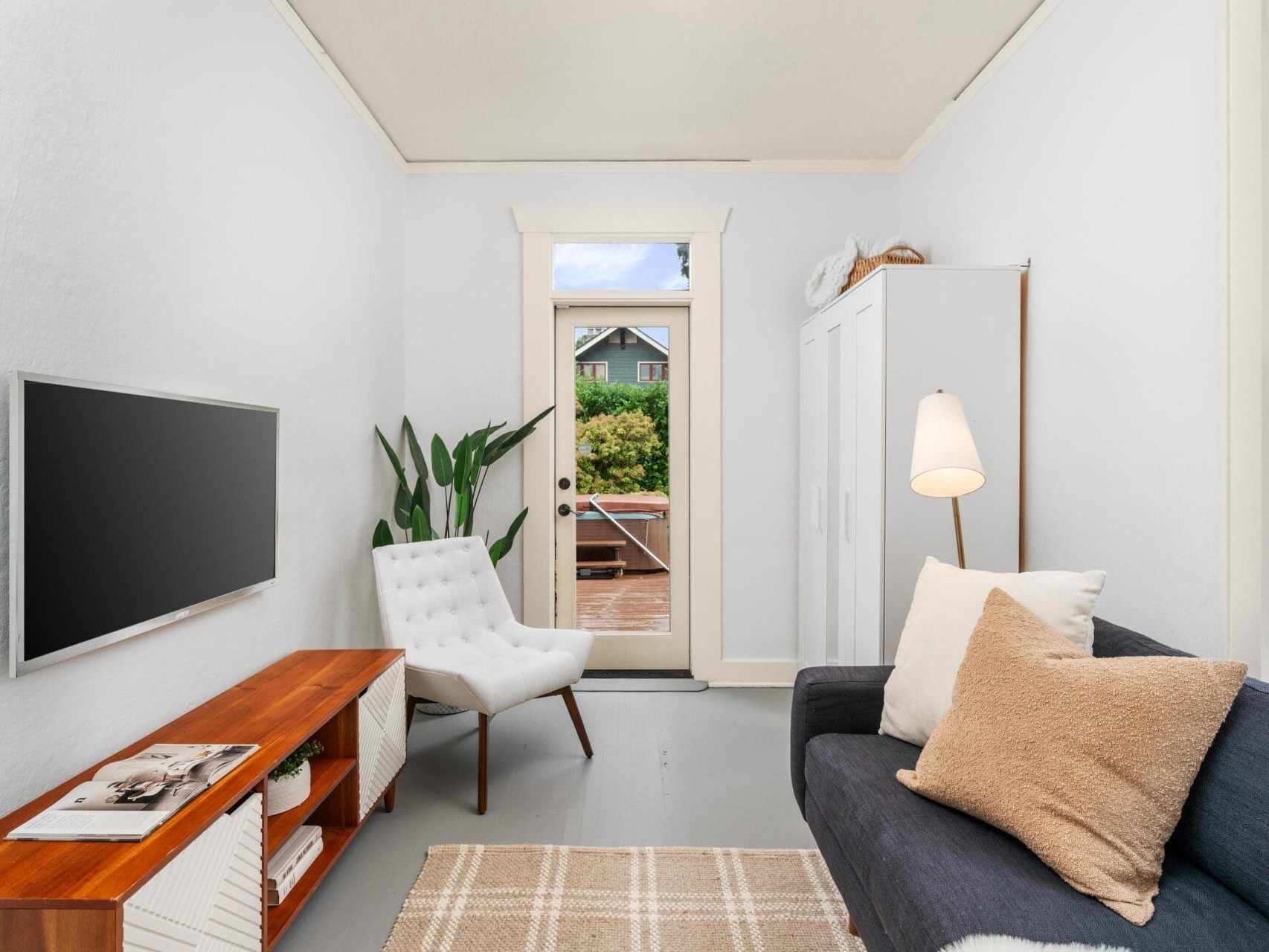 A cozy, narrow living room with a white armchair, dark sofa, and a wall-mounted TV. A wooden cabinet holds decor, and a potted plant stands beside the armchair. A door with a window leads outside. Soft lighting and neutral tones create a calm ambiance.