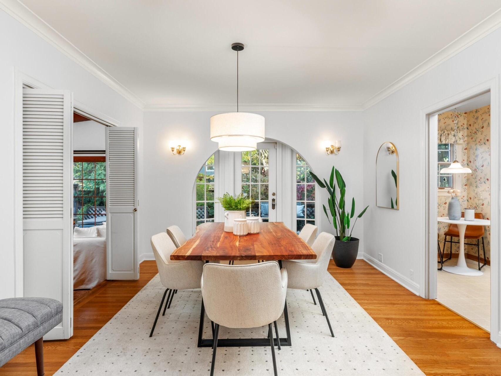 A bright dining room with a wooden table, cream chairs, and a large pendant light. A potted plant stands by a curved window that lets in natural light. An adjoining room with floral wallpaper is visible. Neutral tones create a cozy atmosphere.