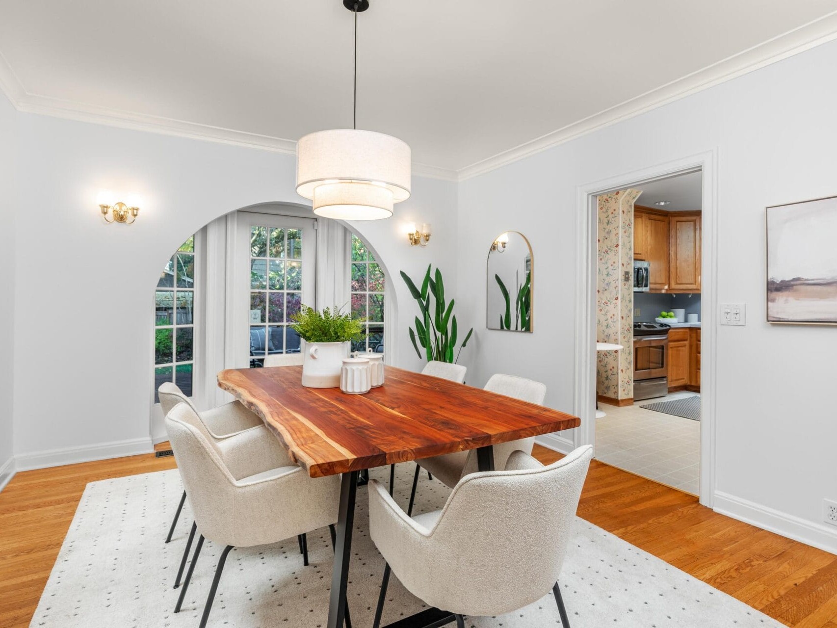 A modern dining room with a wooden table and six white upholstered chairs. A large pendant light hangs above the table. The room features an arched doorway, potted plants, and a doorway leading to a kitchen. A window showcases greenery outside.