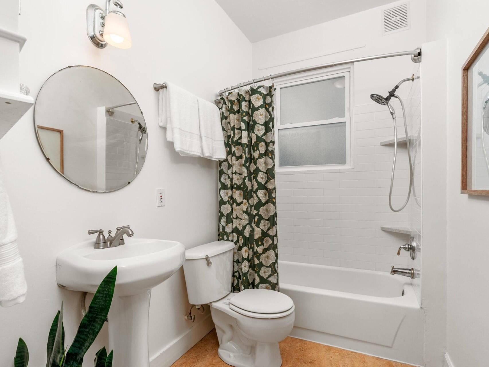 A white bathroom with a round mirror above a sink. Theres a toilet and a bathtub with a floral shower curtain. A potted plant is on the floor. Towels hang on a wall shelf, and a framed picture is on the right wall.