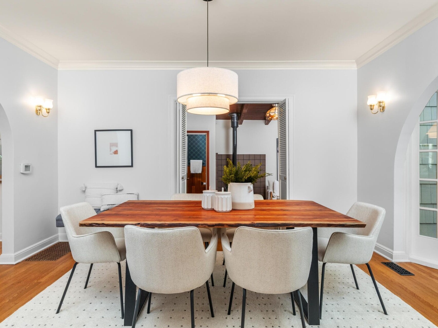 A dining room with a wooden table and eight cushioned chairs. A pendant light hangs above the table, and the walls are painted in a light gray shade. Two arched doorways and framed art are visible, creating an elegant and modern atmosphere.
