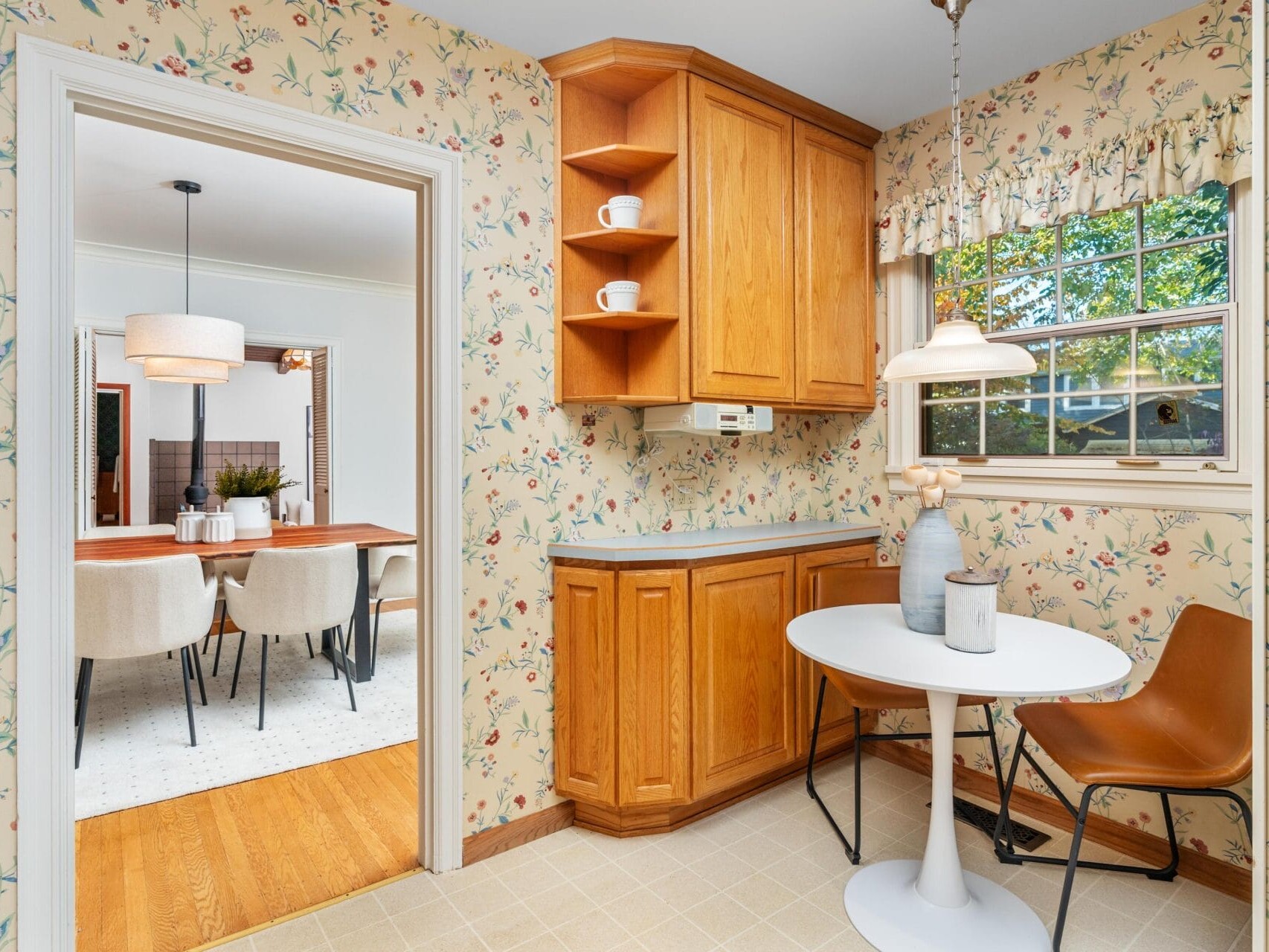 Cozy kitchen corner with floral wallpaper, featuring a small round table, a brown chair, wooden cabinets, and a window with a patterned valance. The adjoining dining room has a wooden table and beige chairs, visible through an open doorway.
