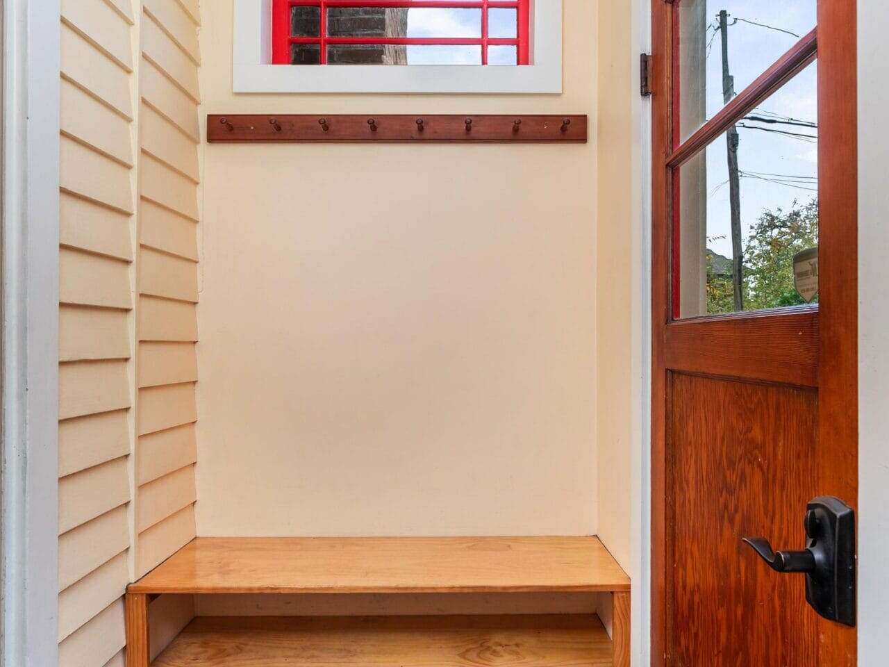 A small entryway with a wooden bench and shoe storage underneath. A wooden door with a window is on the right, and a window above the bench has red trim. A row of hooks for hanging coats is mounted on the wall.