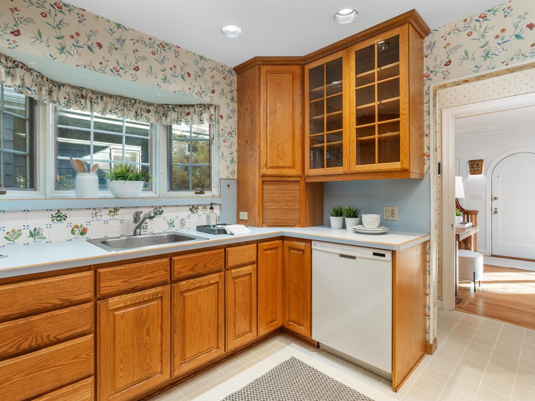 A cozy kitchen with wooden cabinets and floral wallpaper. It features a corner sink, dishwasher, and large windows with decorative curtains. A small rug is on the floor, and two small potted plants sit on the countertop.