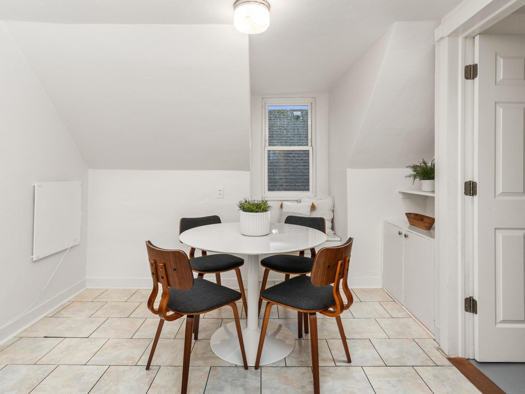 A cozy dining area featuring a round white table with four wooden chairs. A small window is centered on the back wall, and theres a plant in the middle of the table. The walls and floor are light-colored, with a white door on the right.