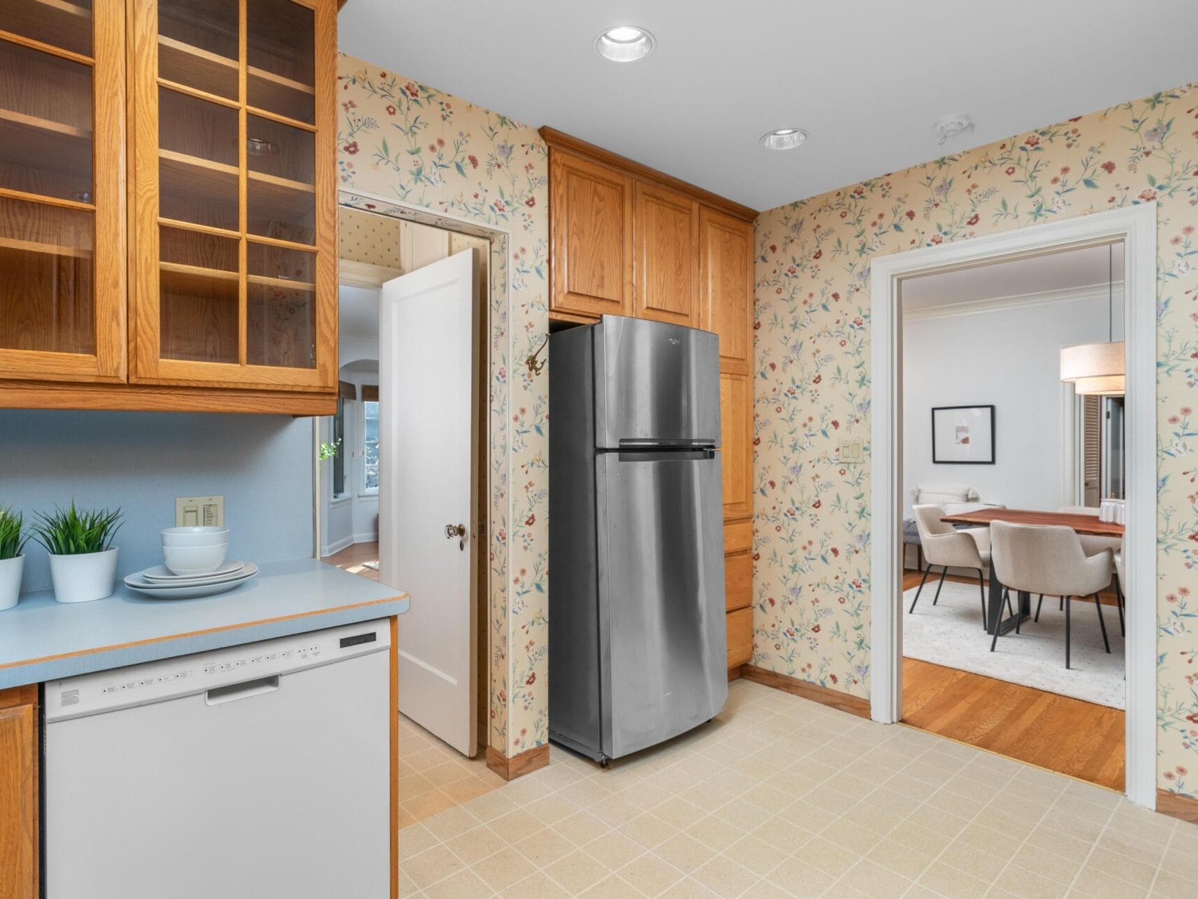A kitchen with floral wallpaper, wooden cabinets, and a stainless steel refrigerator. A dishwasher is next to a counter with potted plants. Two doorways lead to other rooms, one with a dining table and chairs.