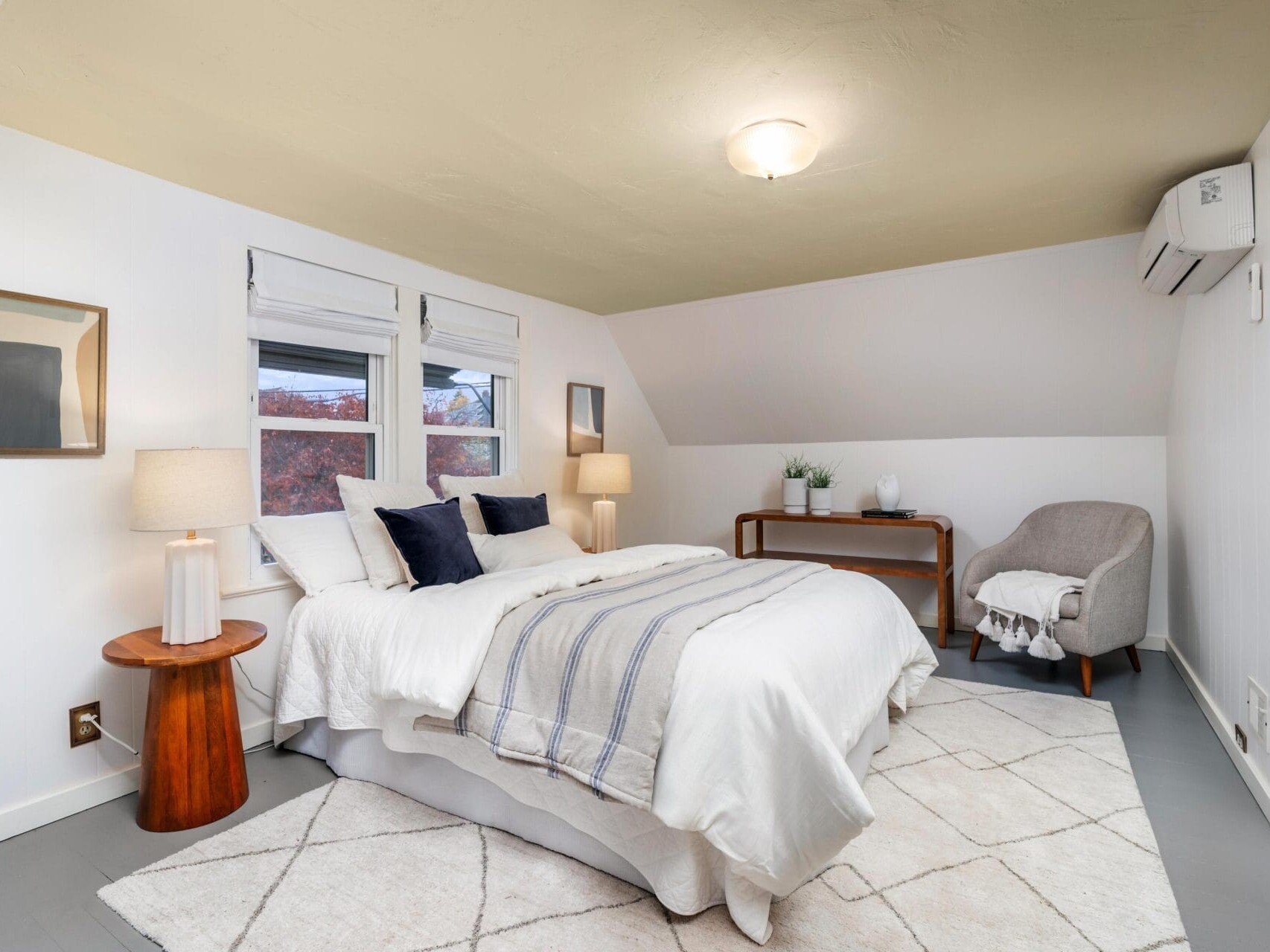 A cozy bedroom features a neatly made bed with white and striped linens, two bedside tables with lamps, a small desk with a plant, a cushioned chair with a throw, and minimalistic decor on white walls. A soft, patterned rug covers the floor.