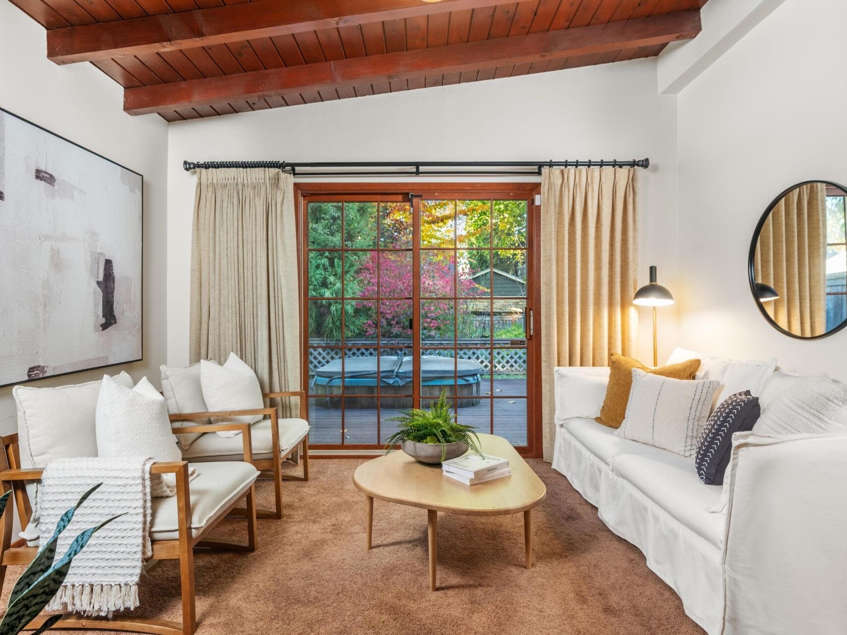 A cozy living room with a white sofa, two wooden chairs with white cushions, and a round wooden table. A large abstract painting and a round mirror decorate the walls. Glass doors reveal a patio with greenery outside. Soft, natural lighting brightens the space.