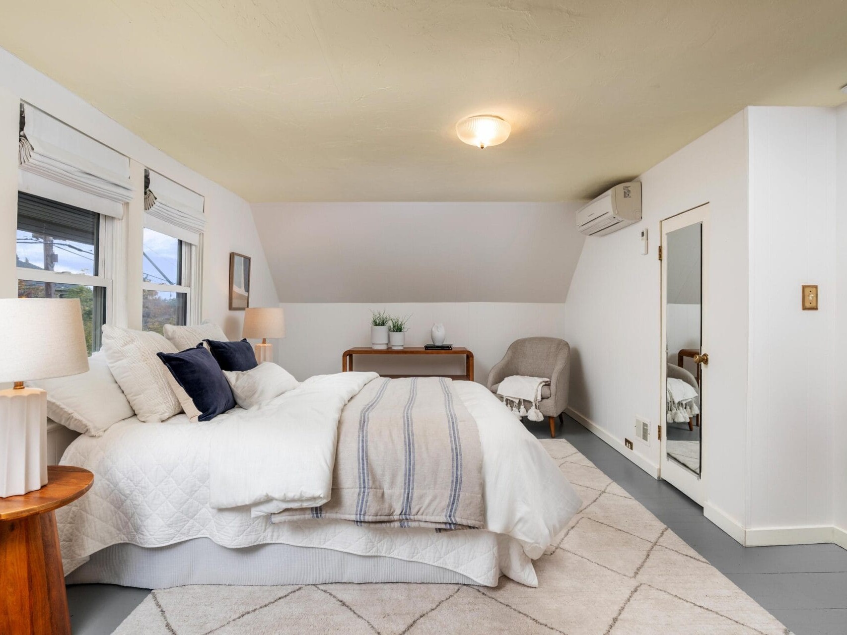 A cozy bedroom with a king-sized bed, white and gray bedding, wooden side tables, and beige carpeting. A small armchair sits near a large mirror. A fire extinguisher is mounted on the wall, and a small window allows natural light.