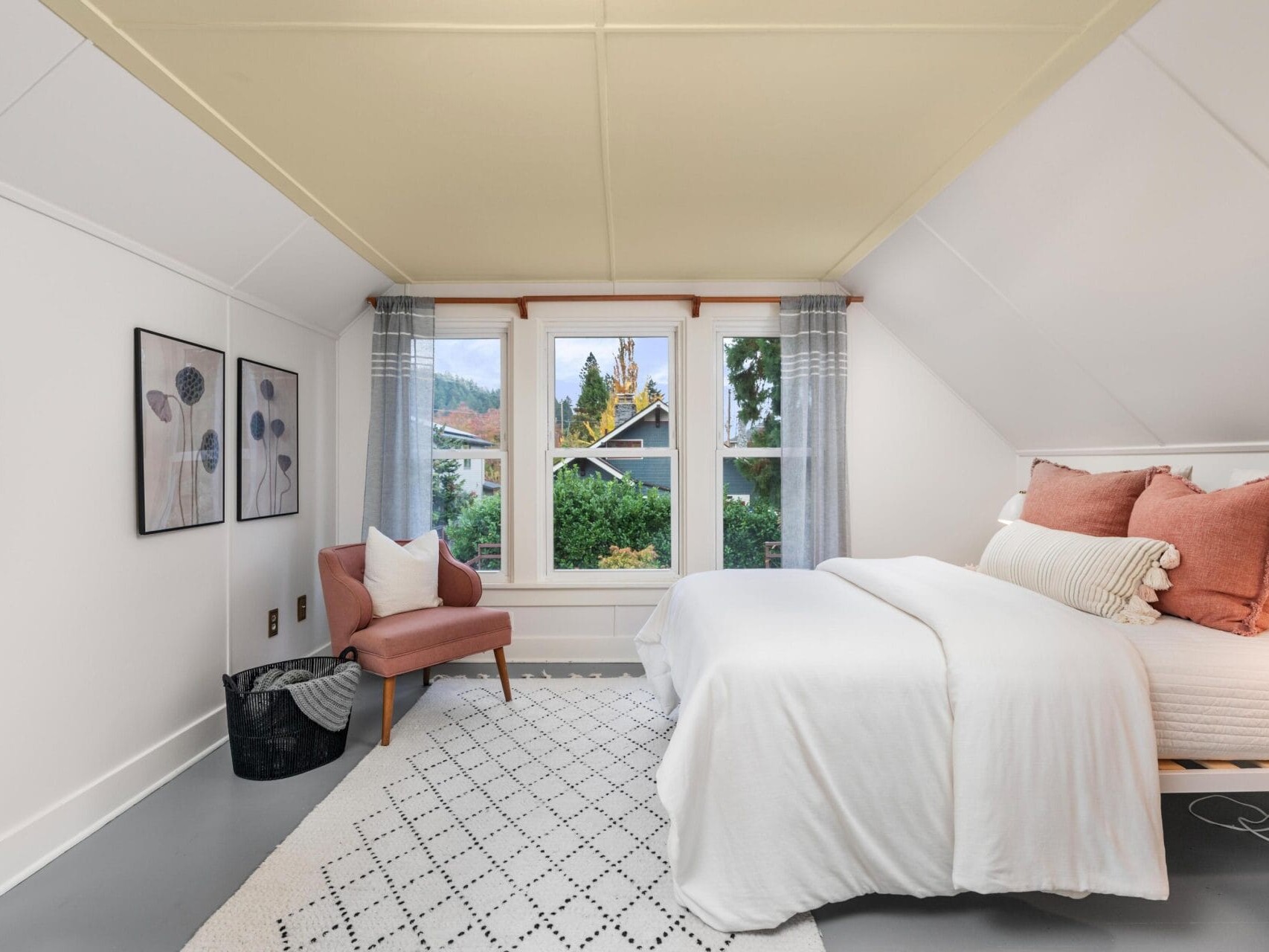 A cozy bedroom with white walls and a sloped ceiling. It features a bed with white and peach bedding, a peach armchair, a patterned rug, and three framed pictures on the wall. Large windows offer a view of greenery outside.