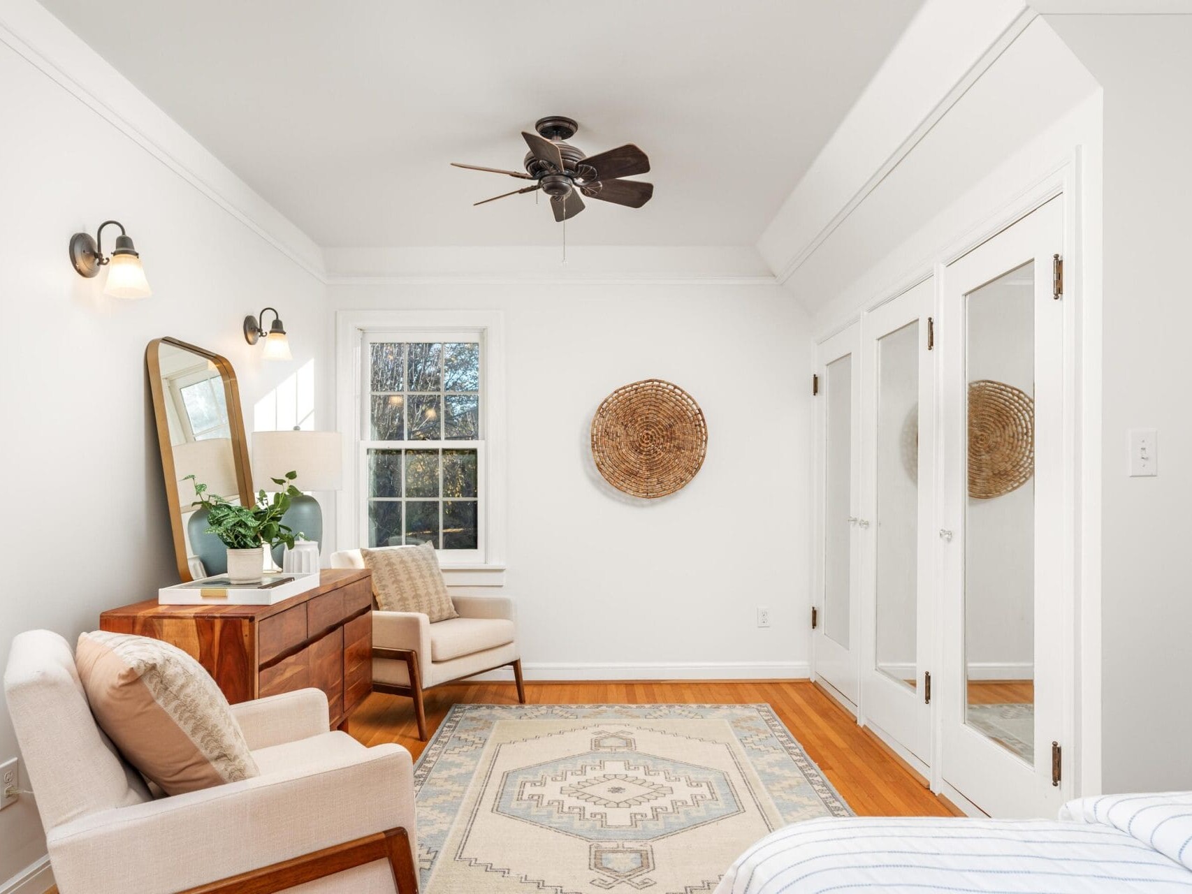 A bright bedroom with white walls and hardwood floors. It features a ceiling fan, two beige armchairs, a wooden dresser with a mirror, a potted plant, a woven wall decoration, and a large area rug. Natural light enters through a window.