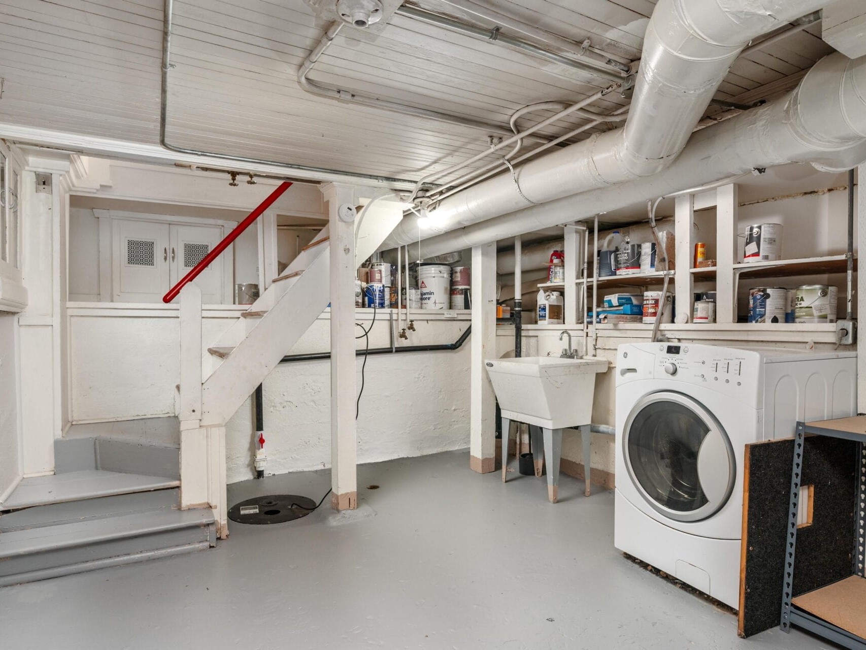 A basement laundry room with a washing machine beside a utility sink. Shelves above the sink hold various paint cans and supplies. Stairs with a red railing lead to an upper floor. Exposed pipes run along the ceiling and walls.