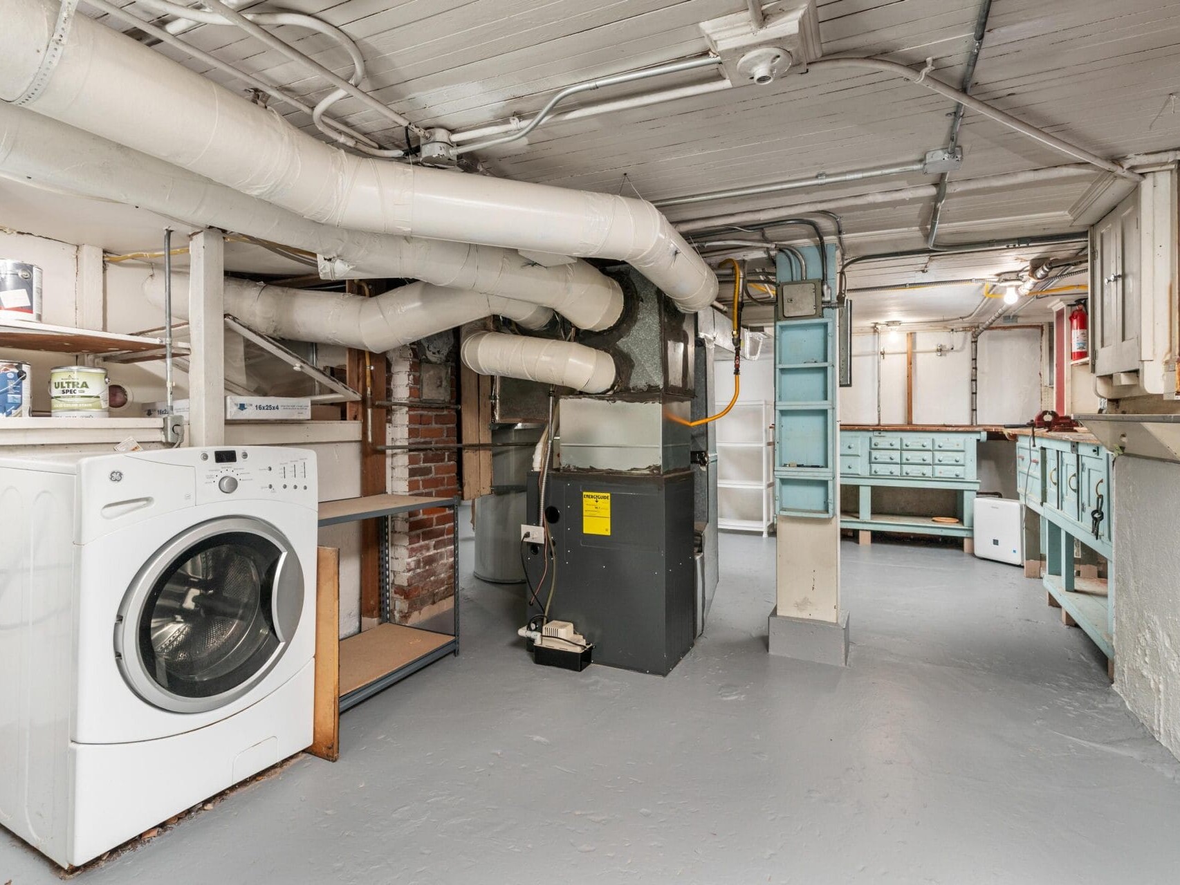 A basement with exposed pipes on the ceiling, featuring a white front-loading washing machine on the left. Shelving and drawers line the walls, with a large HVAC system in the center. The floor is painted a light gray.