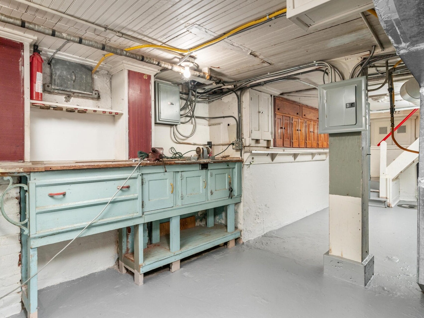 A basement workshop with a light blue workbench, wooden cabinets, electrical panels, and overhead wiring. The walls and floor are painted white, and a fire extinguisher is mounted on the wall. Stairs lead up on the right.