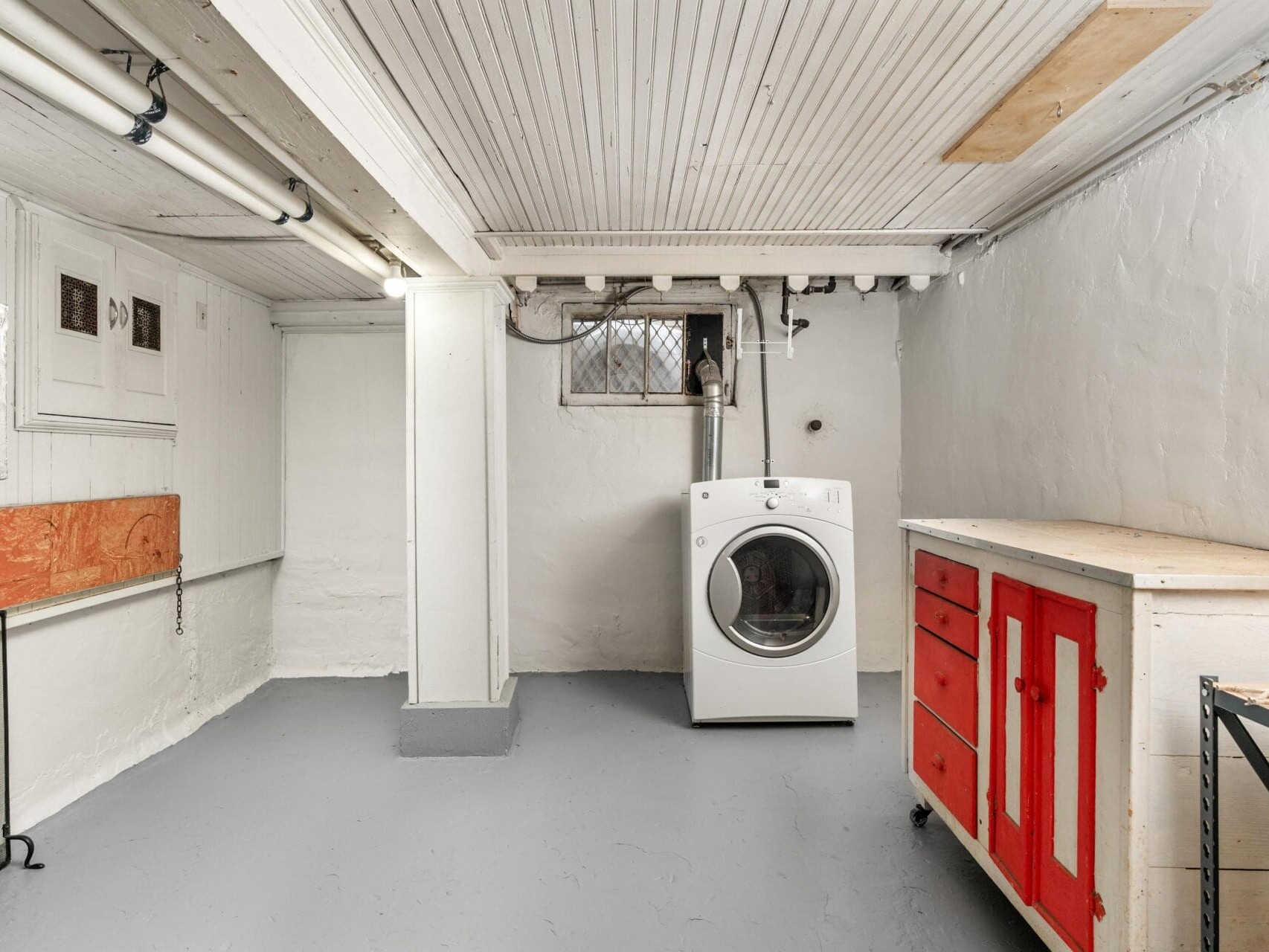 A basement laundry room with white walls and exposed pipes. There is a washing machine centered against the wall, a wooden workbench with red drawers on the right, and small windows near the ceiling. The floor is painted gray.