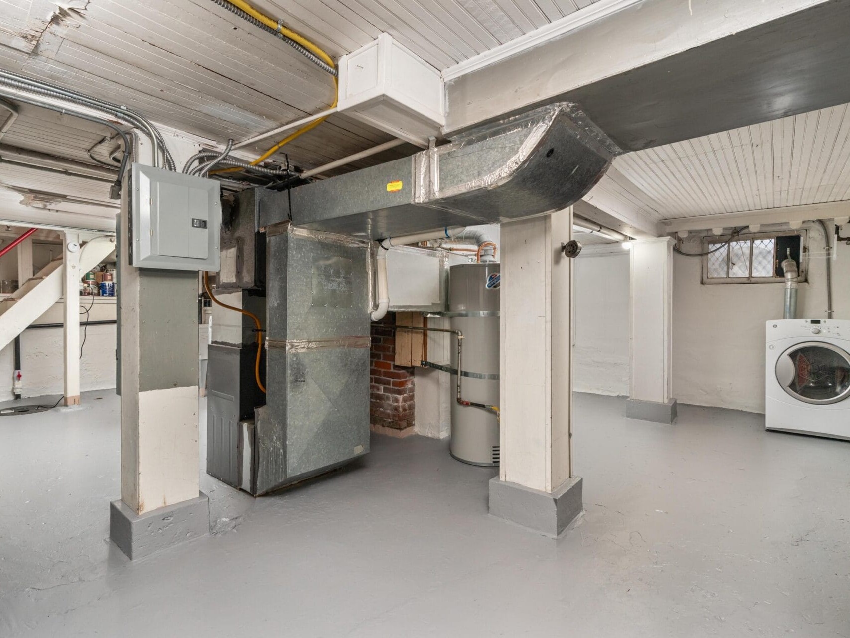 A clean basement with exposed pipes and beams. In the center, theres a furnace and a water heater. To the right, a washing machine is placed against the wall. The floor and ceiling are painted light gray.