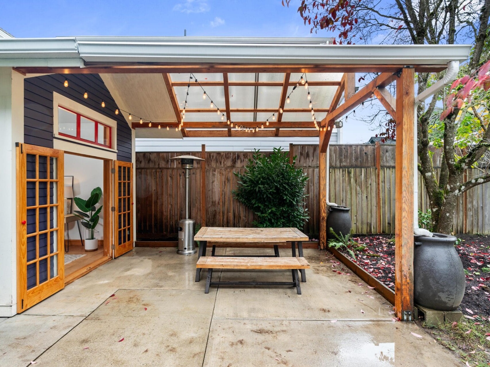 Outdoor patio with a wooden pergola, string lights, and a picnic table. The space is adjacent to a blue building with open French doors. A small potted plant and a tall heater are nearby, with a tree and fence in the background.