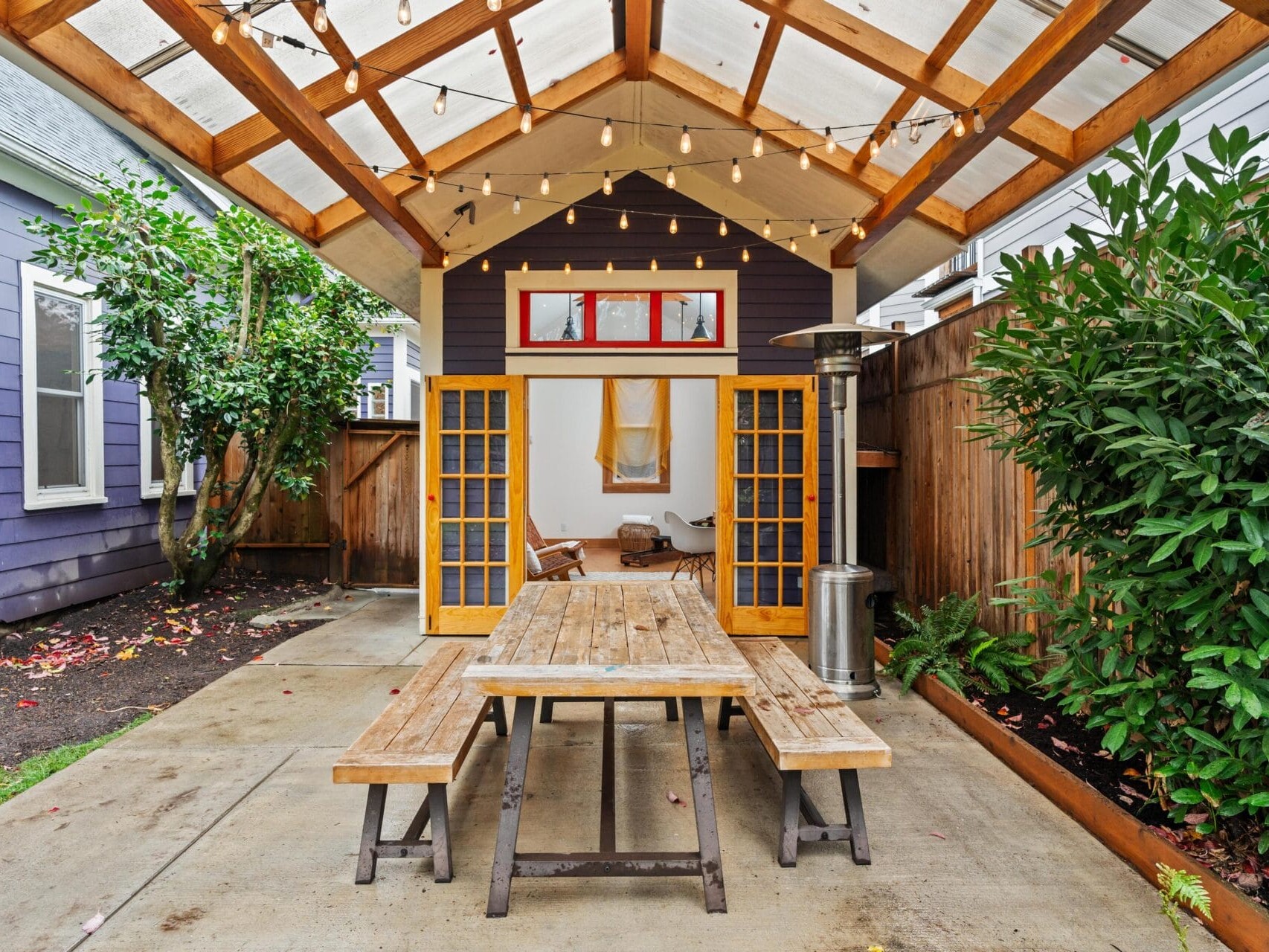 A cozy outdoor patio features a wooden picnic table under a wooden pergola adorned with string lights. The area is enclosed by a wooden fence and has a small building with open doors, showcasing a seating area inside. Shrubs and plants line the sides.