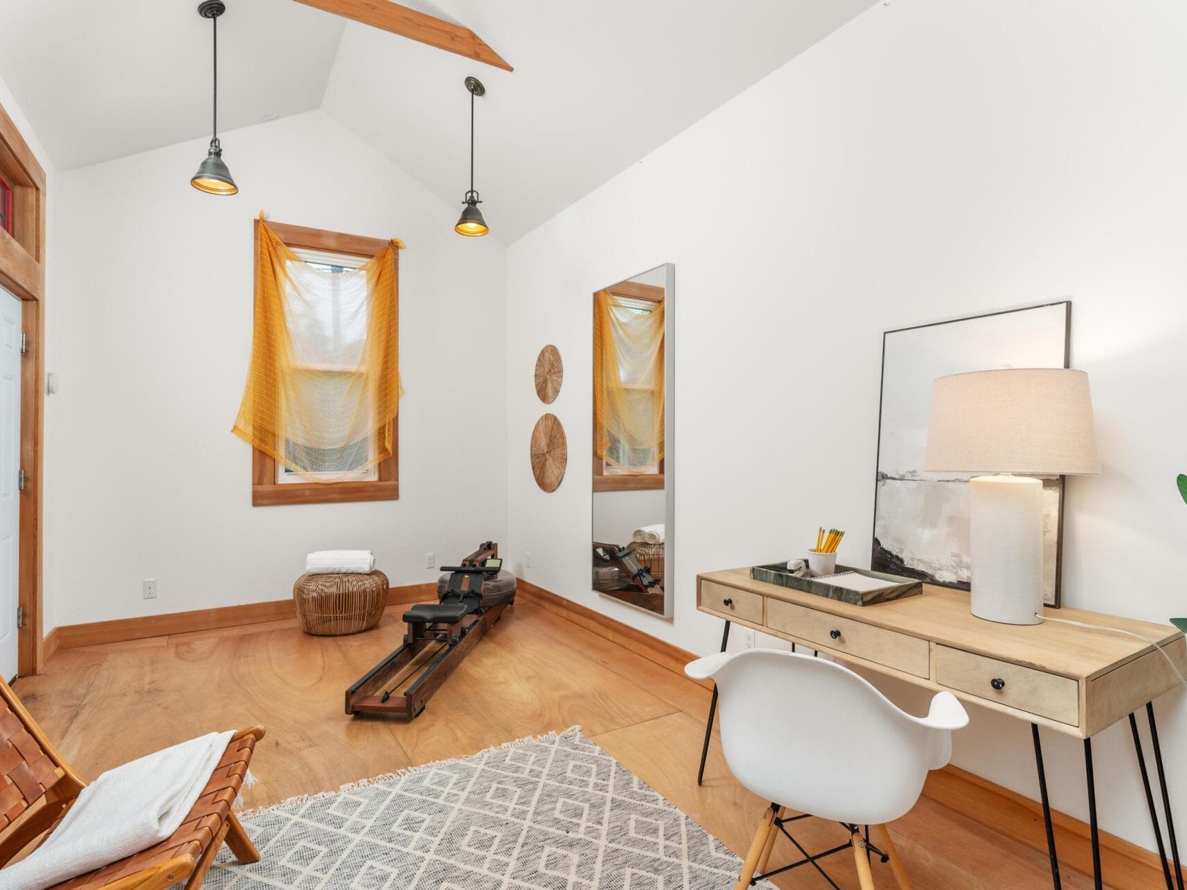 A minimalist exercise room with a rowing machine, a wooden floor, and a woven rug. A desk with a chair, lamp, and decor accents the corner. Warm lighting and yellow curtains add a cozy touch to the space.