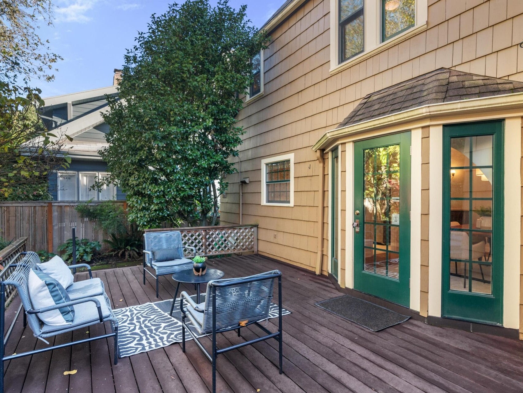 A cozy backyard deck featuring a seating area with blue cushioned chairs around a small table on a patterned rug. The deck is attached to a beige house with green-framed double doors and surrounded by a wooden fence and lush greenery.