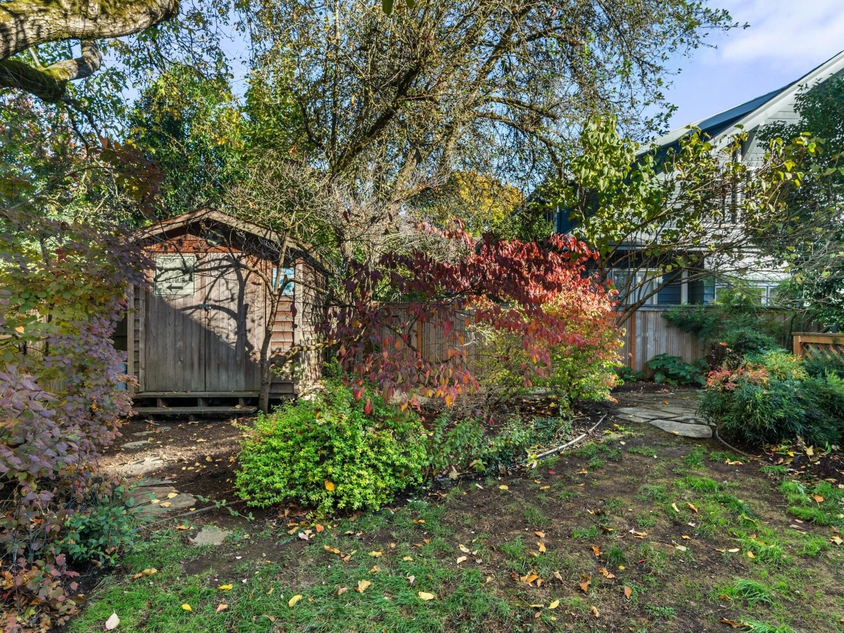 A lush backyard features a wooden shed on the left, surrounded by vibrant autumn foliage in red, orange, and green. A pathway leads to the right, with a house partially visible in the background. Sunlight filters through the trees.