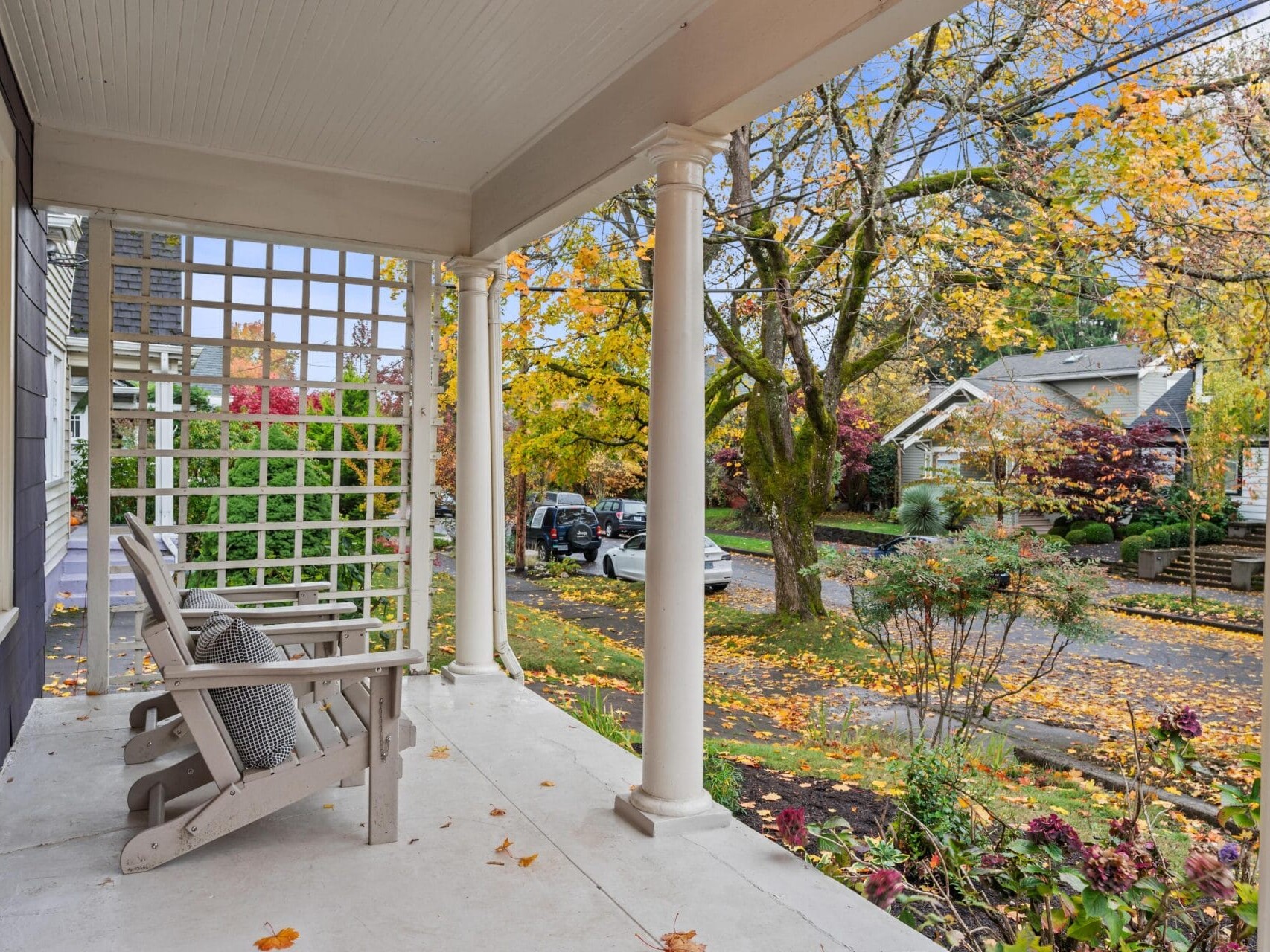 A charming front porch with wooden chairs overlooks a residential street lined with autumn trees. Leaves in vibrant shades of yellow and orange cover the ground, while neighboring houses and parked cars are visible in the background.