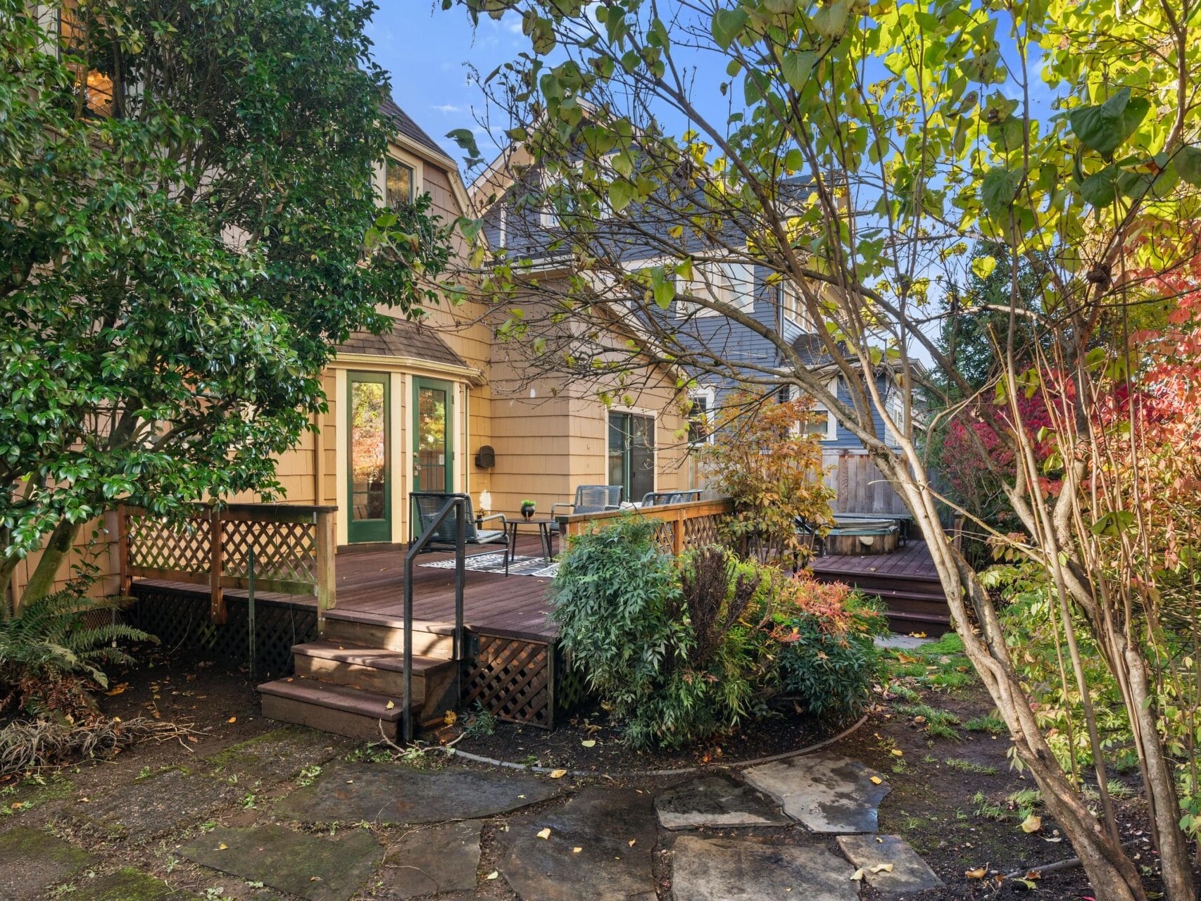A backyard with a wooden deck attached to a house, surrounded by lush green and colorful foliage. The deck includes outdoor seating and a table. Sunlight filters through the trees, creating a peaceful atmosphere.