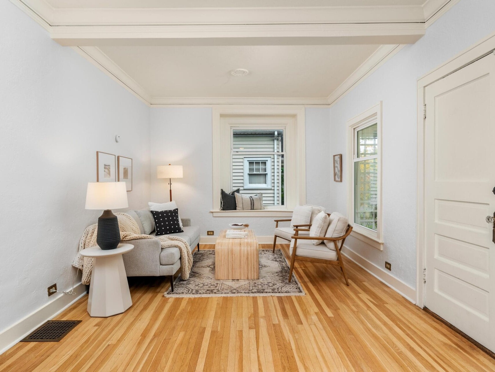 A cozy living room with a small sofa, wooden coffee table, armchair, and standing lamp. Light wood flooring complements the neutral walls. Two framed pictures hang above the sofa. A window and a door are in view.
