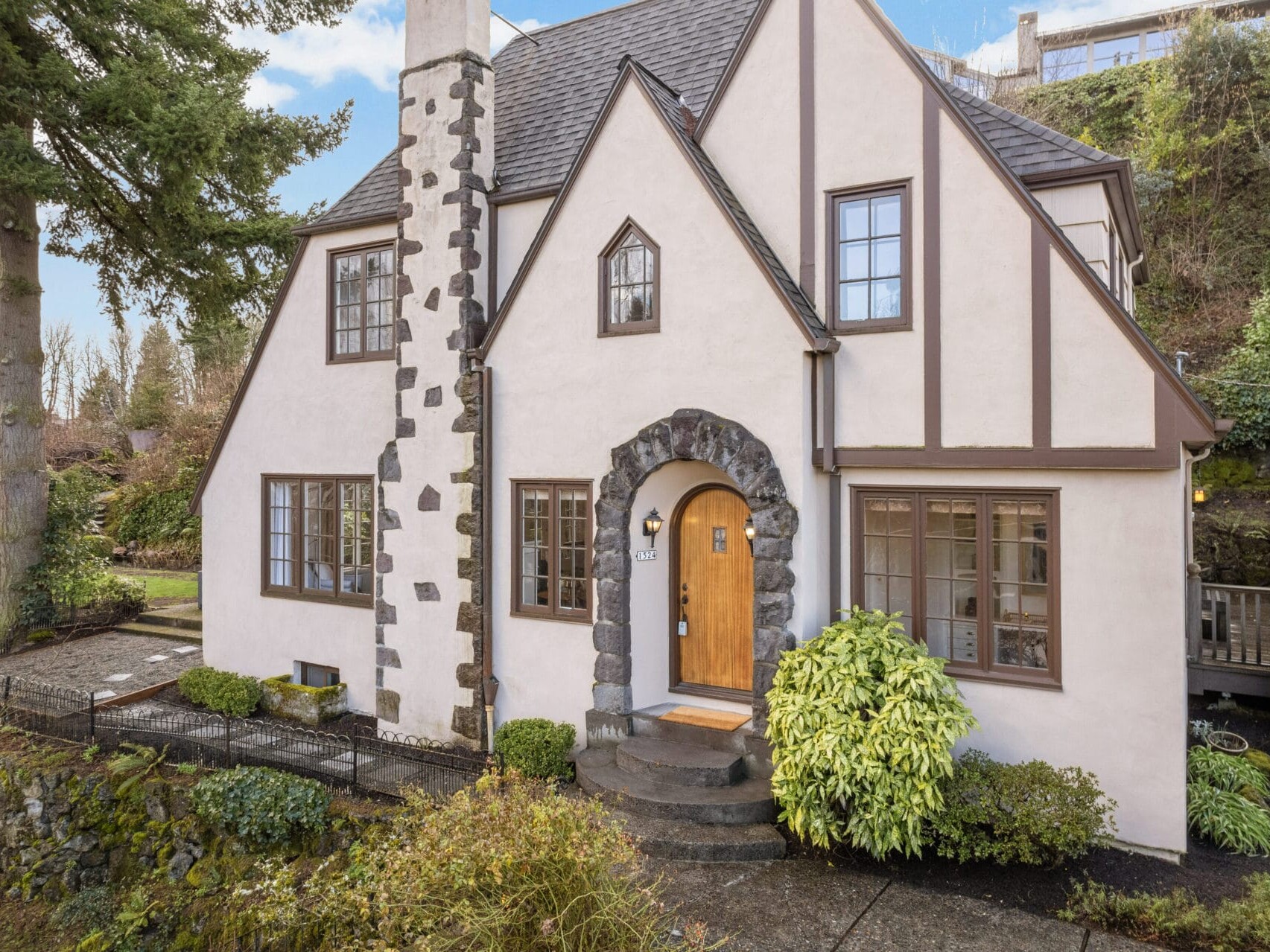 A charming Tudor-style house with a steep roof, stone chimney, and arched wooden door. Surrounded by greenery, it features a garden path and hedges, set against a backdrop of trees and a hill.