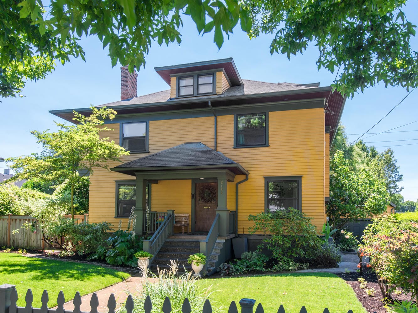 A large, two-story yellow house with gray trim and a chimney, surrounded by lush green trees and bushes. The entrance porch is adorned with plants, and a neatly manicured lawn is visible, enclosed by a wooden picket fence.