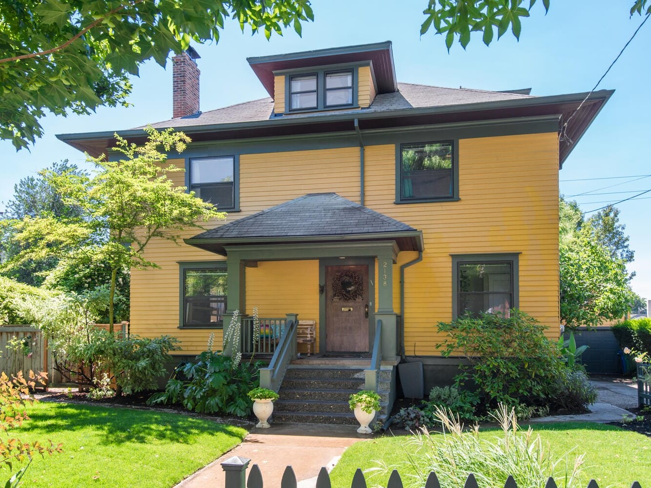 A large, two-story yellow house with a dark roof and green trim is surrounded by lush greenery. The front yard features a small lawn and a path leading to the porch, which is decorated with potted plants. A wooden fence lines the front of the property.