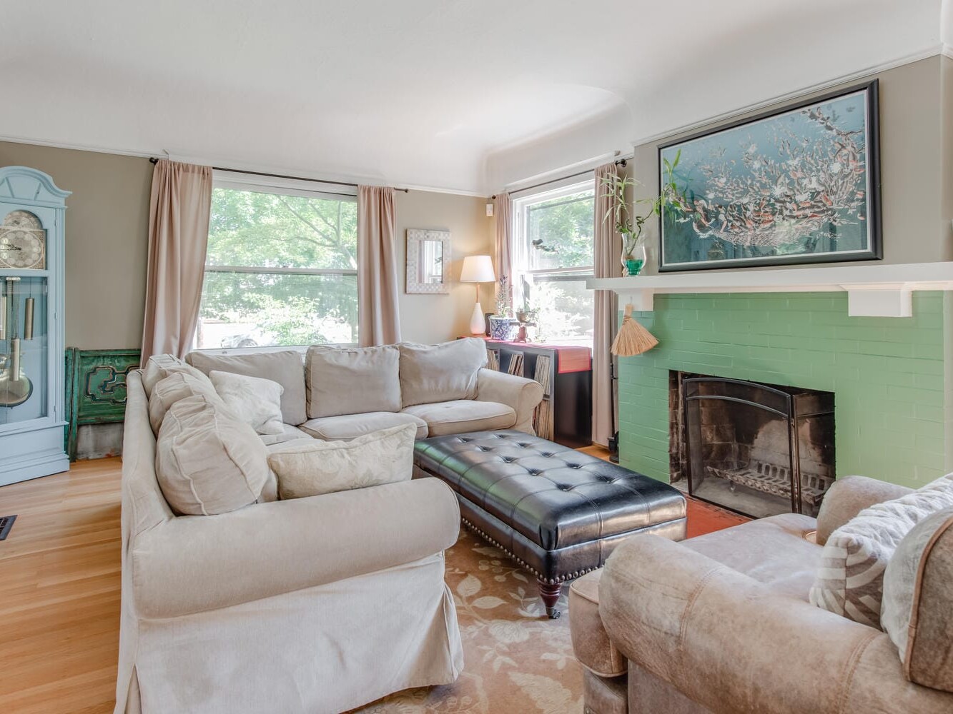 A cozy living room with a large beige sectional sofa, a tufted black ottoman, and a green tiled fireplace with a framed painting above it. Light streams in through large windows with beige curtains. A blue grandfather clock adorns the corner.