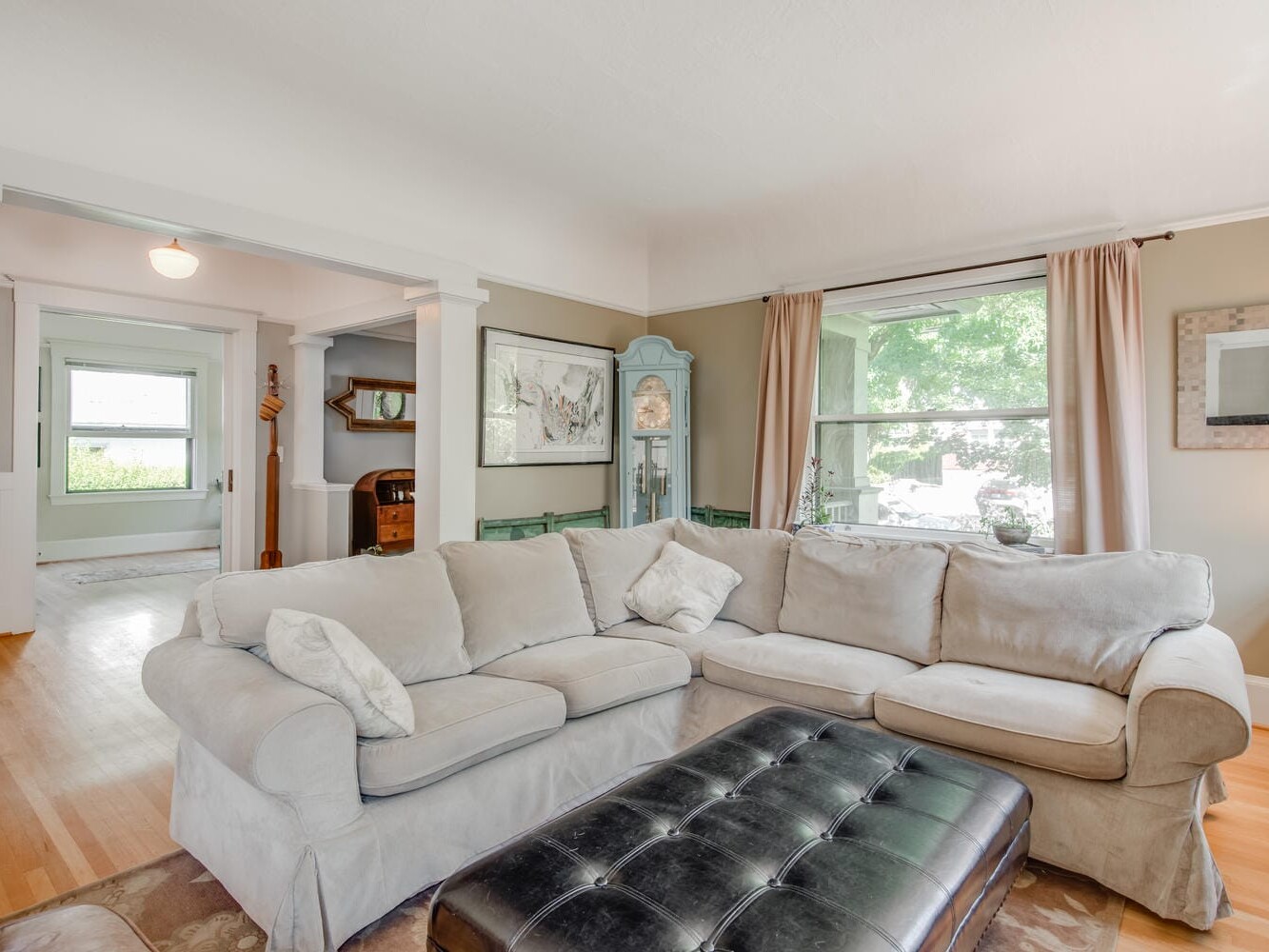 A cozy living room with a beige sectional sofa and a large black ottoman. The room has wooden floors, a large window with curtains, framed artwork, and an open doorway leading to another room with green accents. Natural light fills the space.