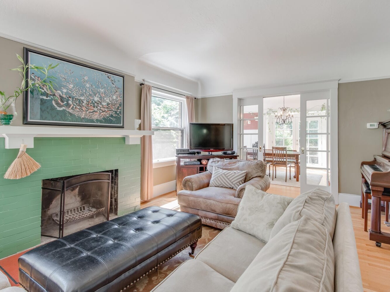 A cozy living room with light-colored sofas and a black ottoman. Theres a green brick fireplace with a large painting above it. A TV sits near a window, and a piano is on the right. Glass doors lead to a dining area with a chandelier.