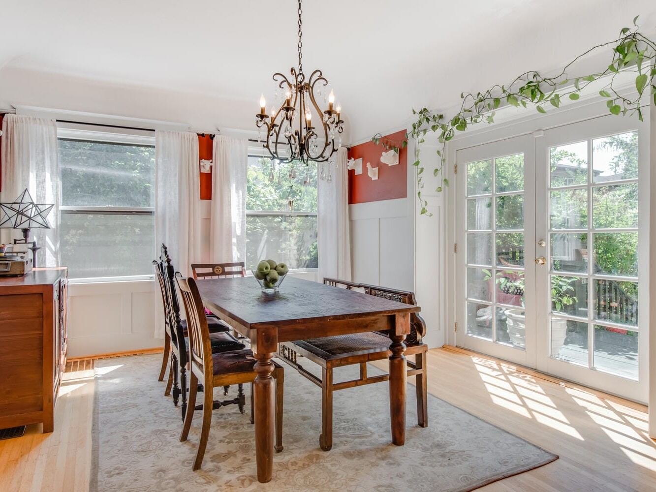 A bright dining room with a wooden table and eight chairs, a decorative chandelier, and large windows. French doors open to a garden. The room has red and white walls, plant decorations, and a soft rug on a wooden floor.