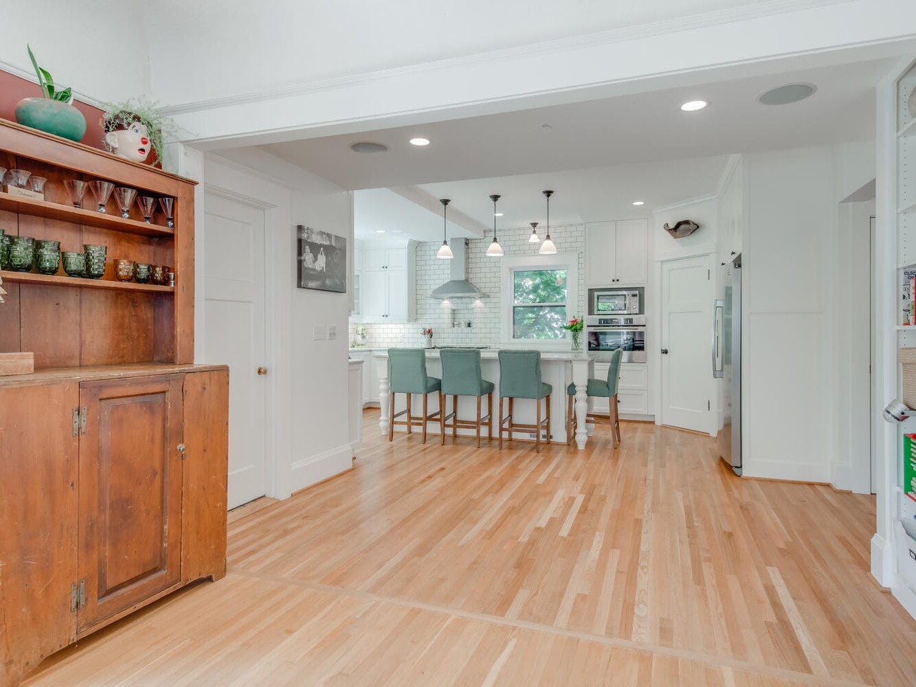 A spacious, modern kitchen with light wood flooring and white walls. It features a wooden cabinet on the left and a large island with turquoise bar stools. Pendant lights hang above the island, and theres a window letting in natural light.
