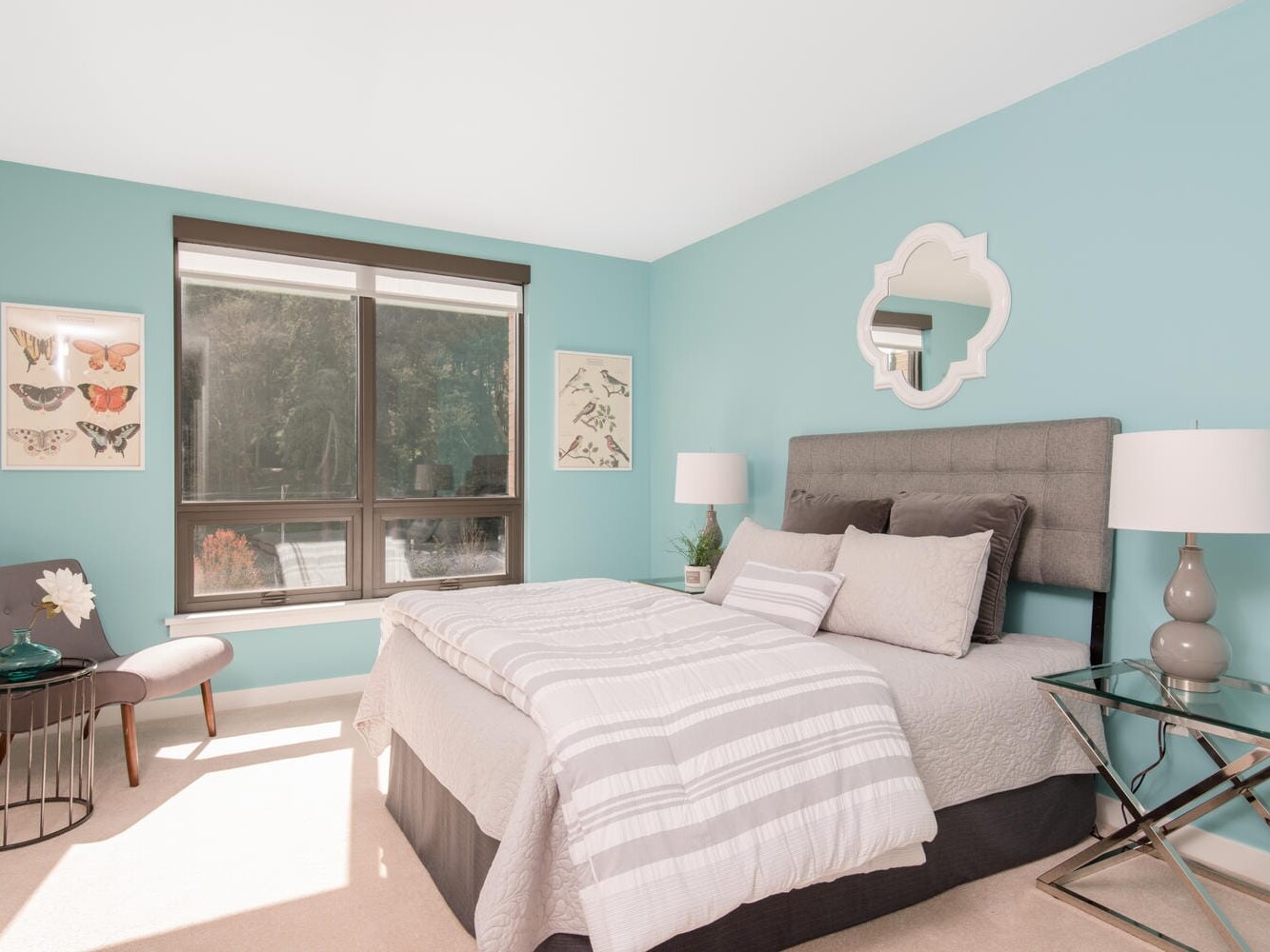 A bedroom with teal walls features a bed with gray and white bedding, two lamps on nightstands, a decorative mirror above the headboard, a chair with a pillow and a flower in a vase, butterfly artwork, and a large window with natural light streaming in.