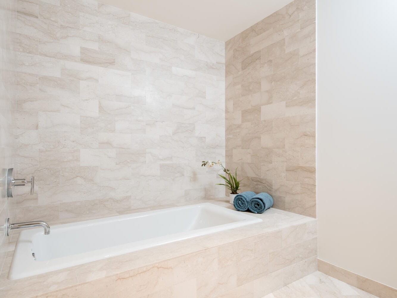 A bathroom featuring a rectangular bathtub with beige stone tiles on the wall and floor. A silver faucet is mounted on the wall. Two rolled blue towels and an orchid in a vase are placed at the corner of the tub.
