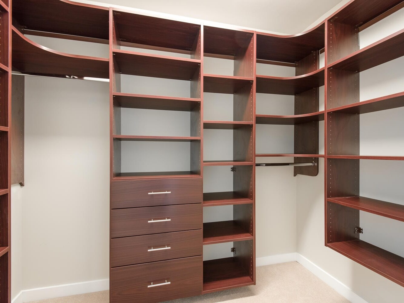 A spacious, empty walk-in closet with dark wood shelving units and drawers against beige walls. The design includes adjustable shelves and hanging rods, providing ample storage space. The floor is carpeted in a light beige color.