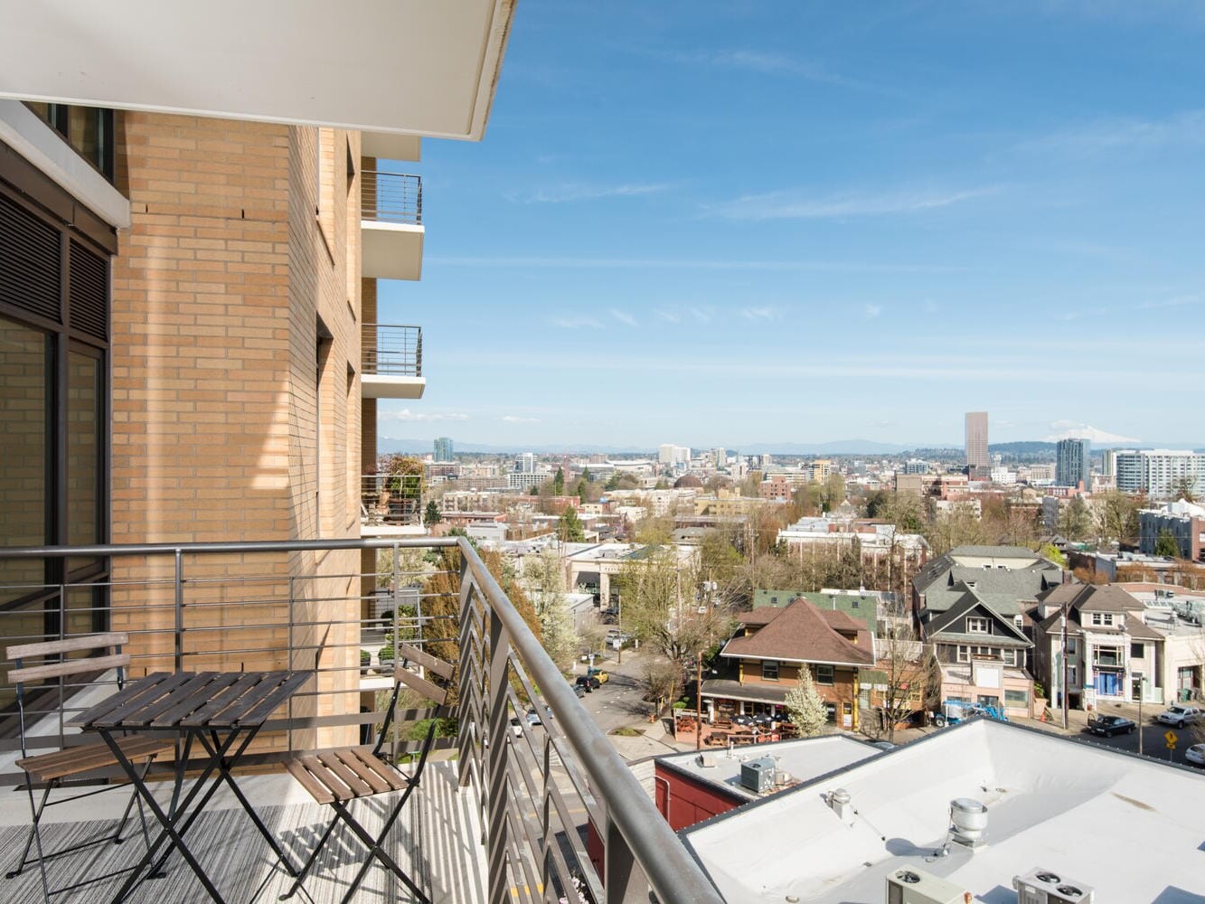 View from a balcony showing a cityscape with a mix of modern and traditional buildings. A small balcony set with two chairs and a table is in the foreground. Mountains and a clear blue sky can be seen in the background.